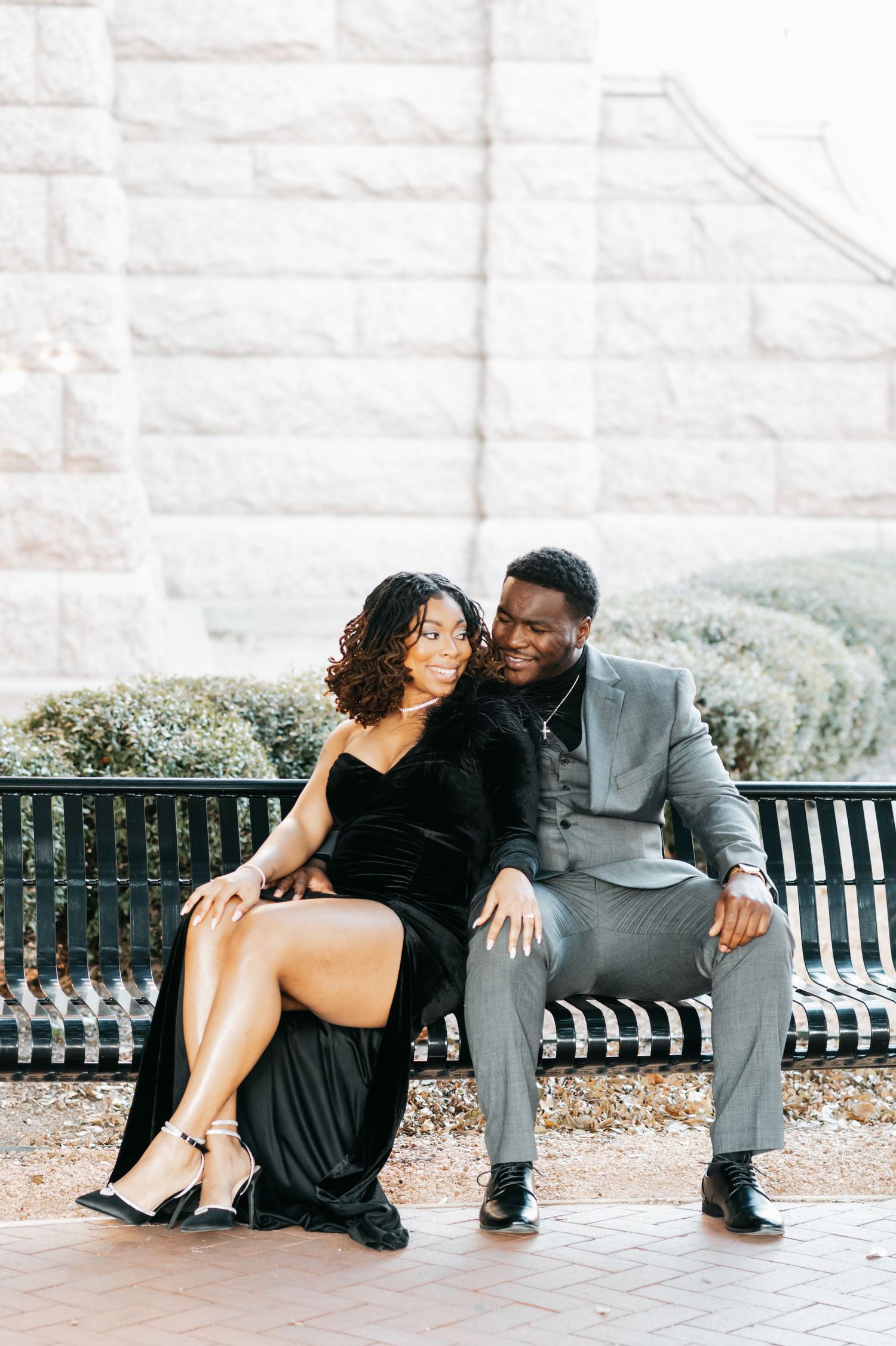 Couple sitting on bench outdoors, woman in black velvet gown, man in gray suit, smiling together.