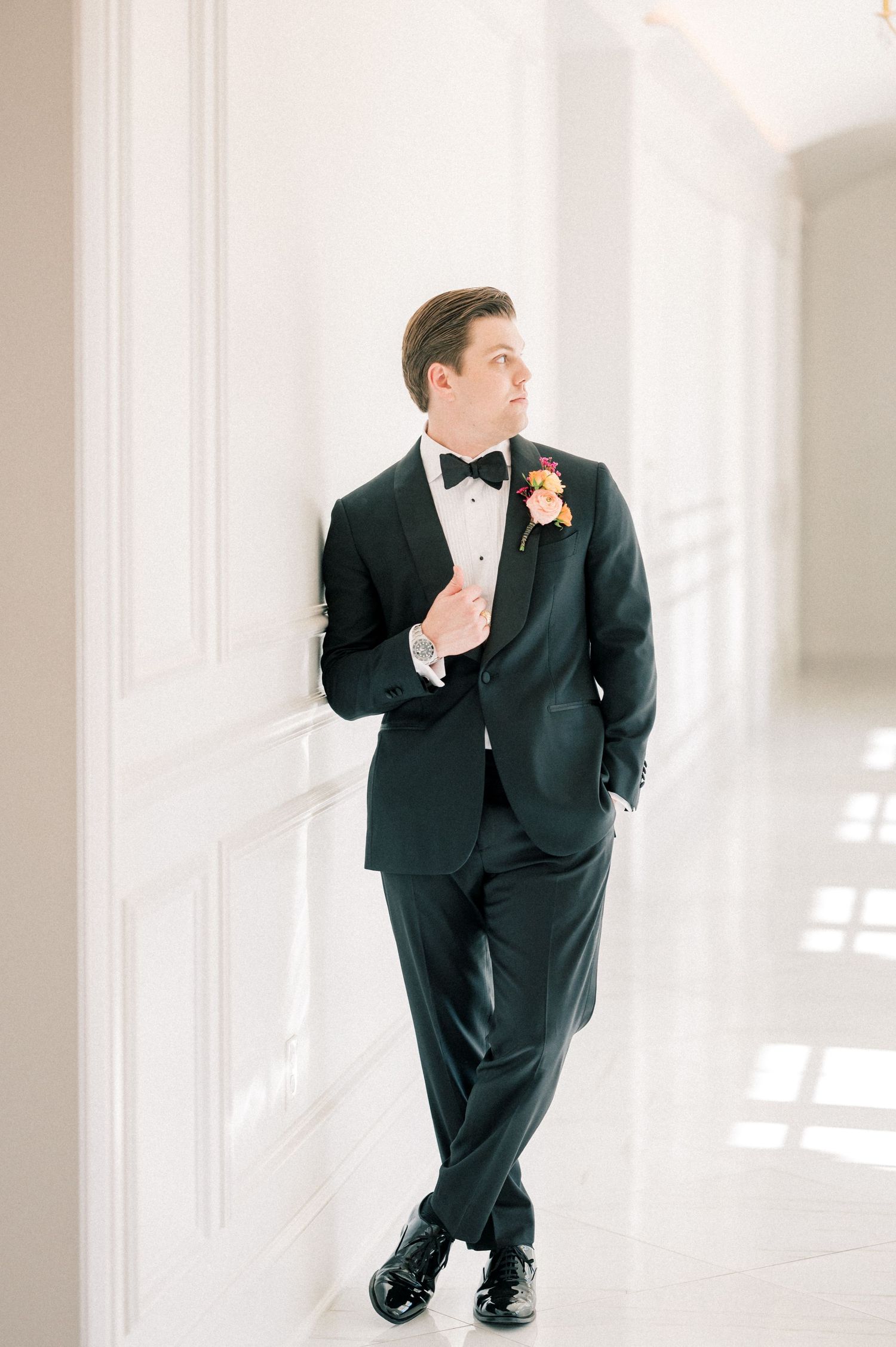 Groom in black tuxedo with bow tie and boutonniere leaning against white wall in elegant venue.