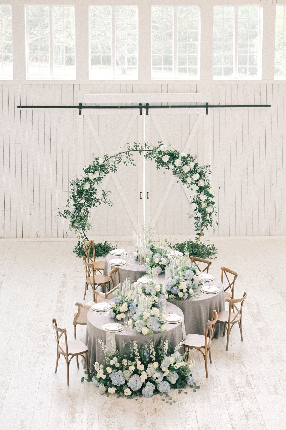 Elegant wedding reception table with gray linen, white floral arch, greenery, and wooden cross-back chairs in a bright white barn.