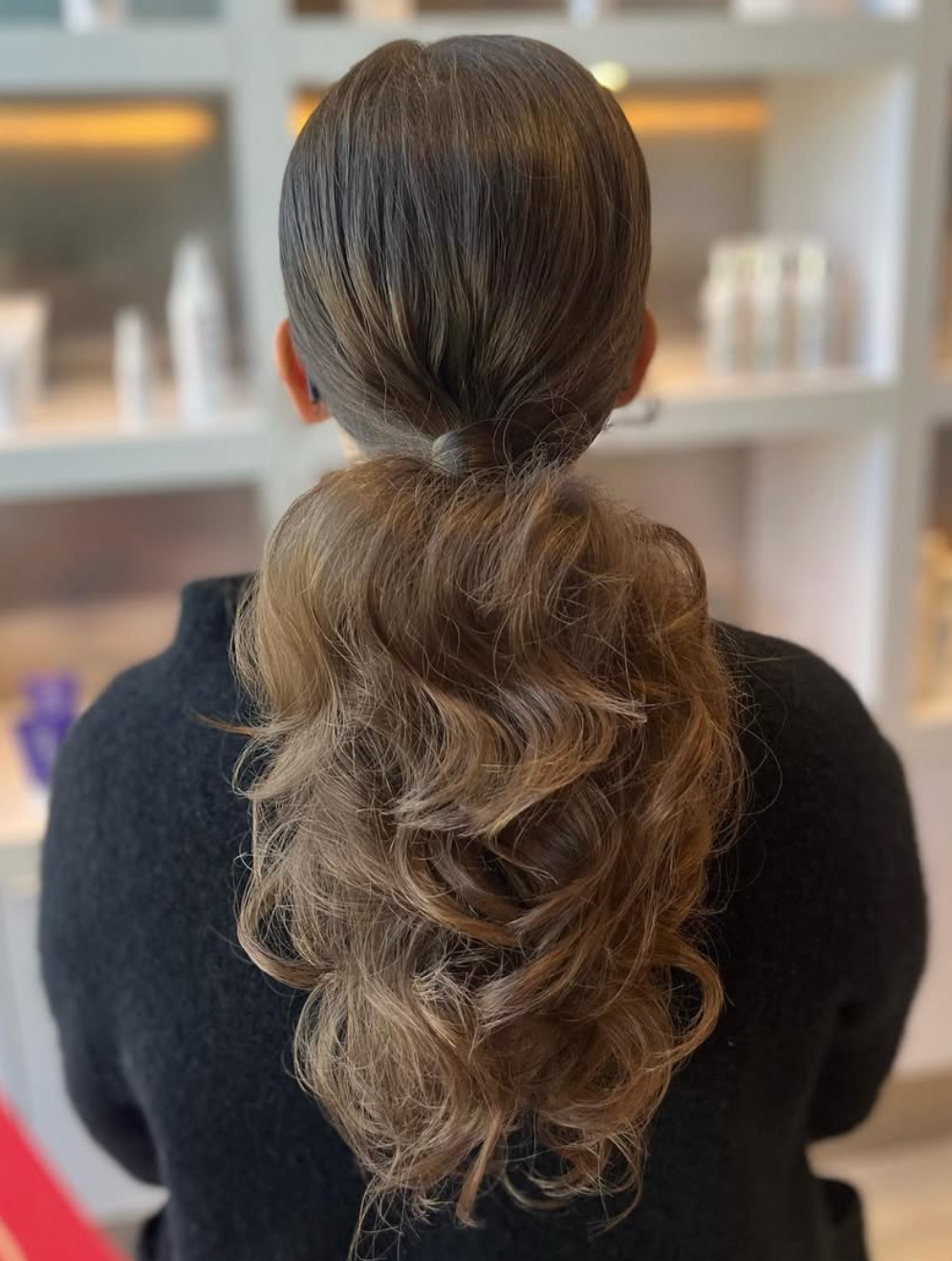 Back view of elegant low ponytail hairstyle with voluminous curls and ombre highlights in a salon setting.