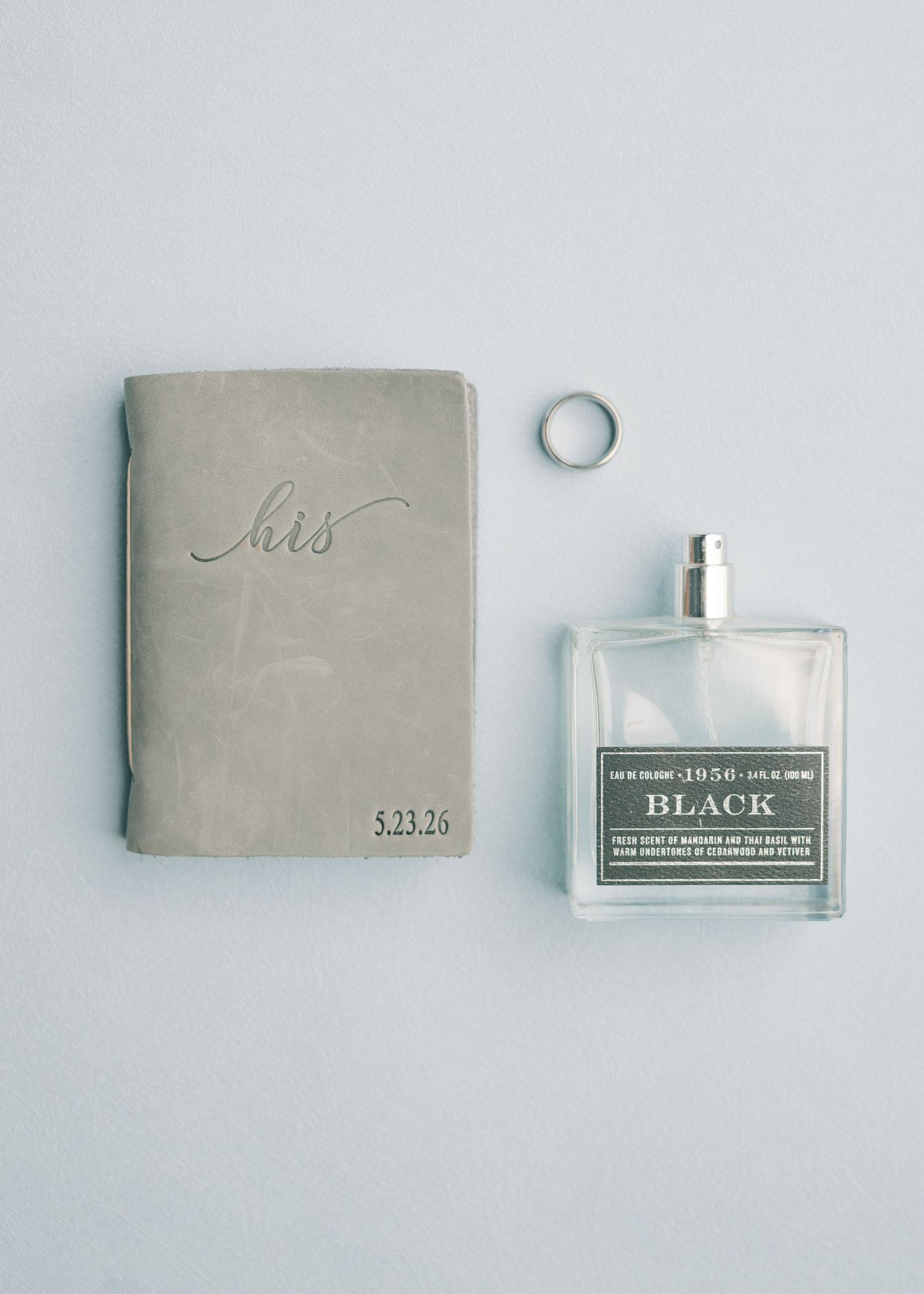 Groom wedding flat lay with his vow book, silver band ring, and Black cologne on blue surface.