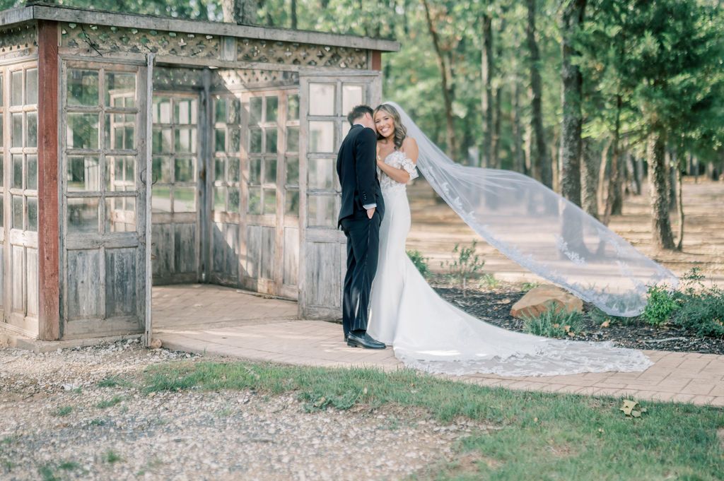 Bride and groom share a romantic embrace on a garden path, with a flowing veil and rustic wooden arbor backdrop.