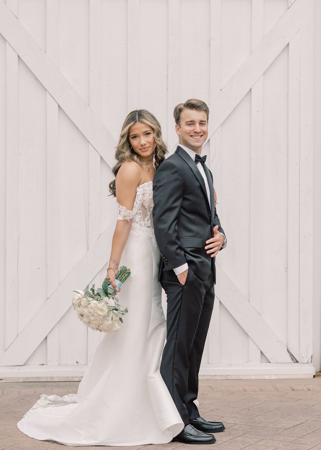 Bride in off-shoulder gown holding bouquet poses back-to-back with groom in black tuxedo before white barn doors.