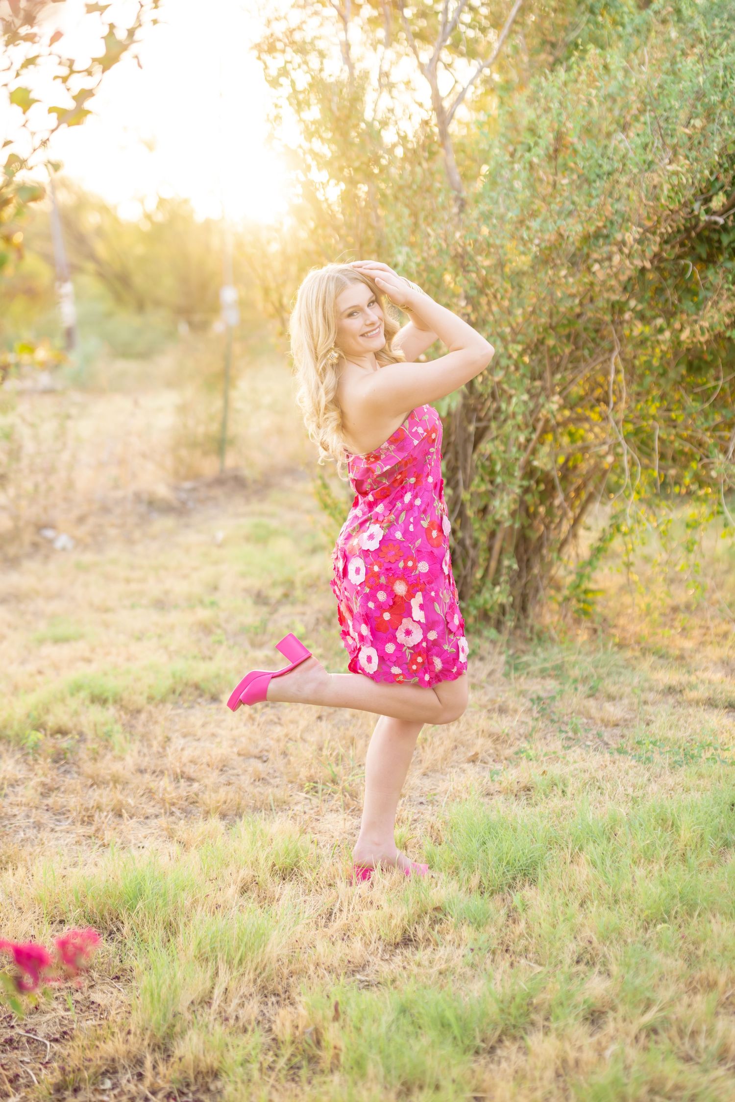 Blonde woman in pink floral sundress posing outdoors in golden sunlight among trees and grass.