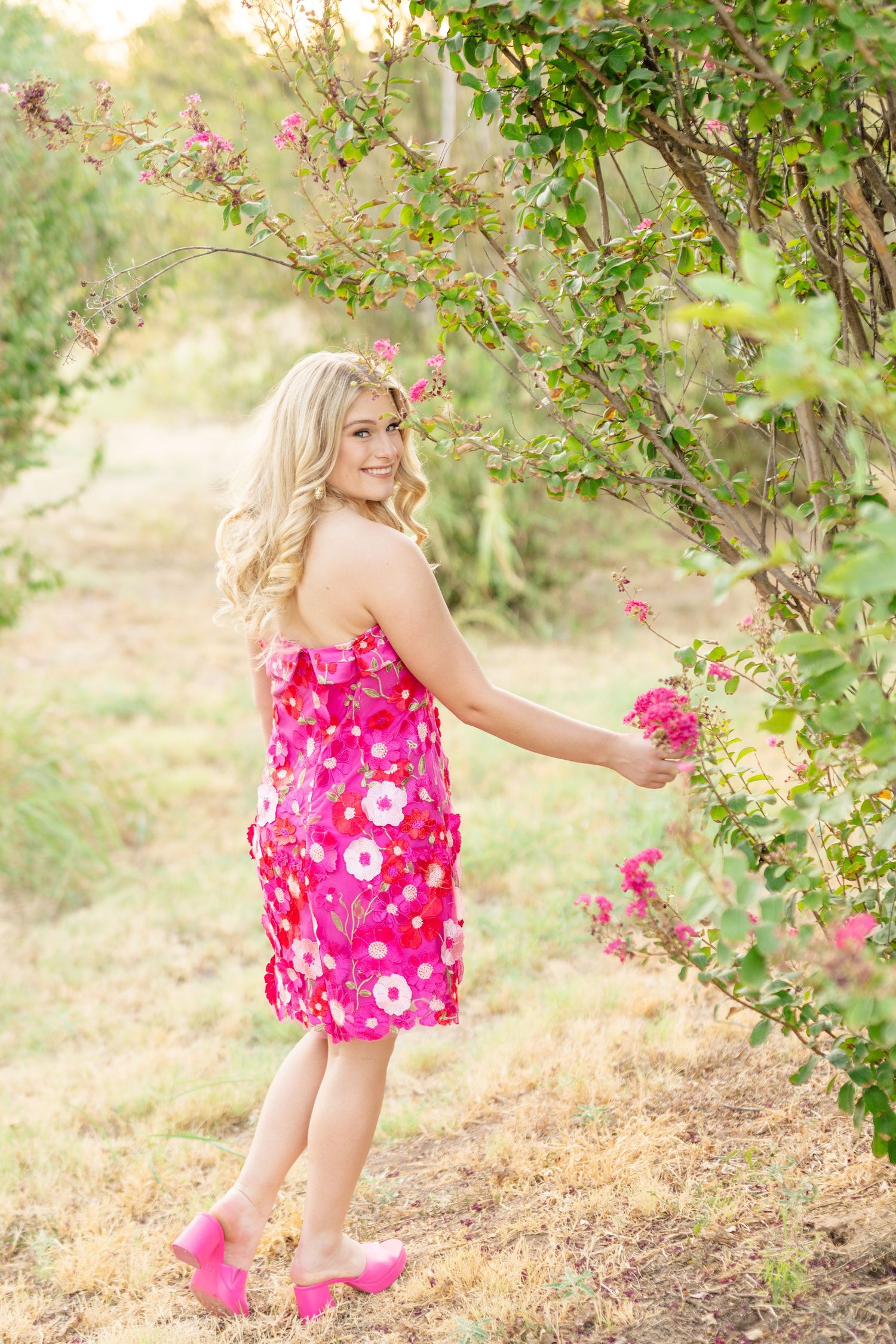 Smiling blonde teen girl in pink floral strapless dress touching blooming pink flowers on outdoor bush.