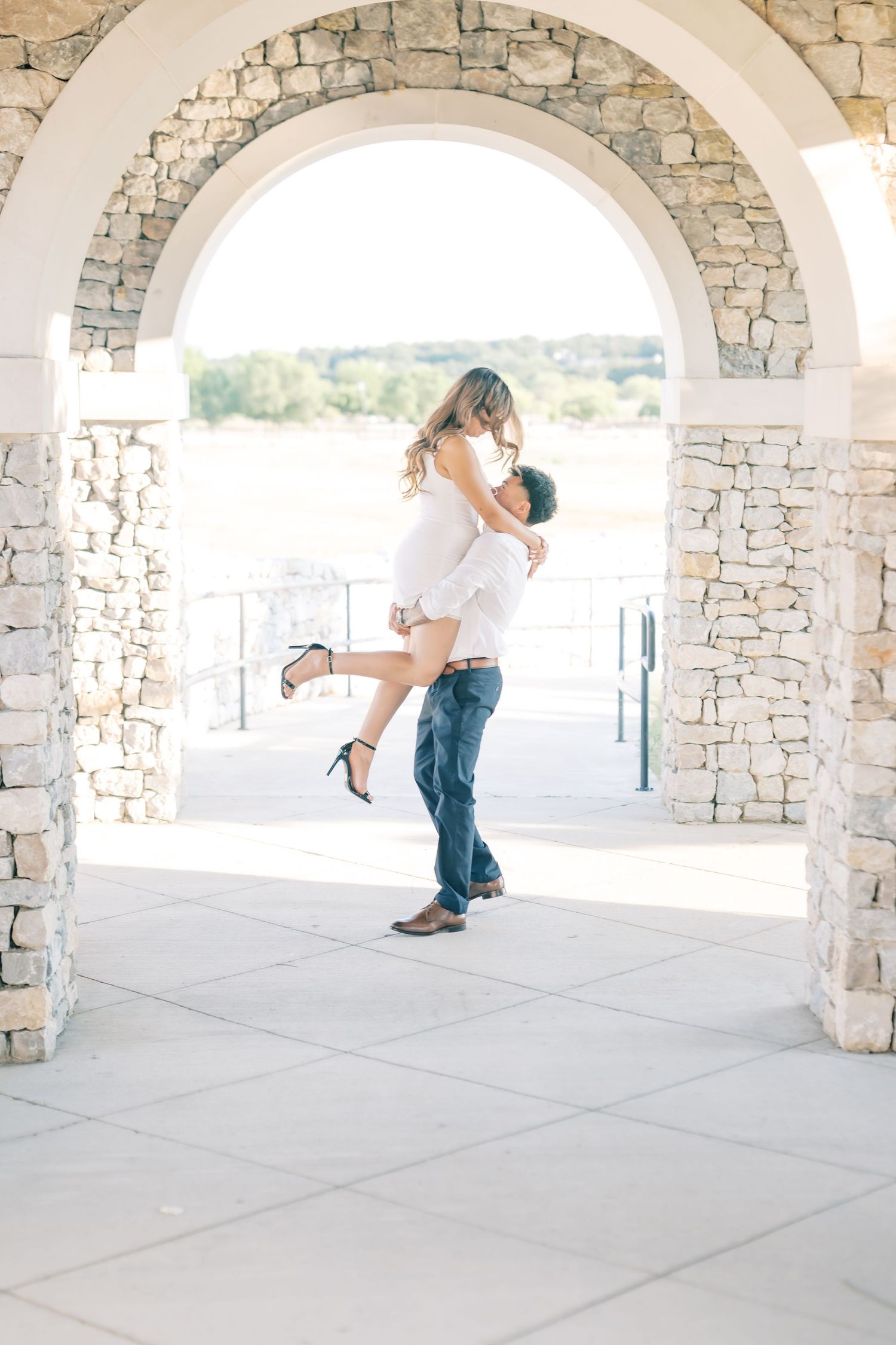 A man lifts his partner in a romantic embrace under a stone arch during an engagement photo session.