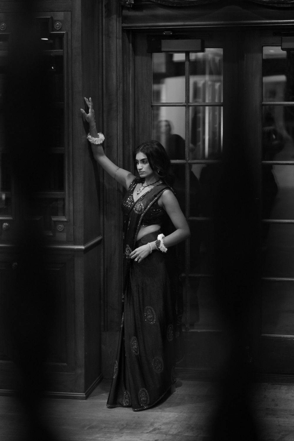 Woman in traditional sari striking dramatic pose against dark doorway in black and white photograph.