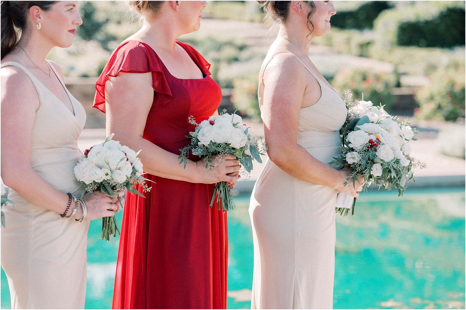 Bridesmaids in red and cream dresses holding white bouquets stand beside a turquoise pool.