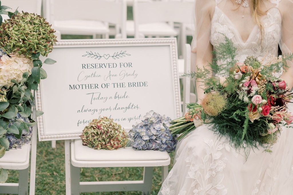 Elegant outdoor wedding ceremony with reserved Mother of the Bride sign, white chairs, hydrangeas, and bride holding colorful bouquet.