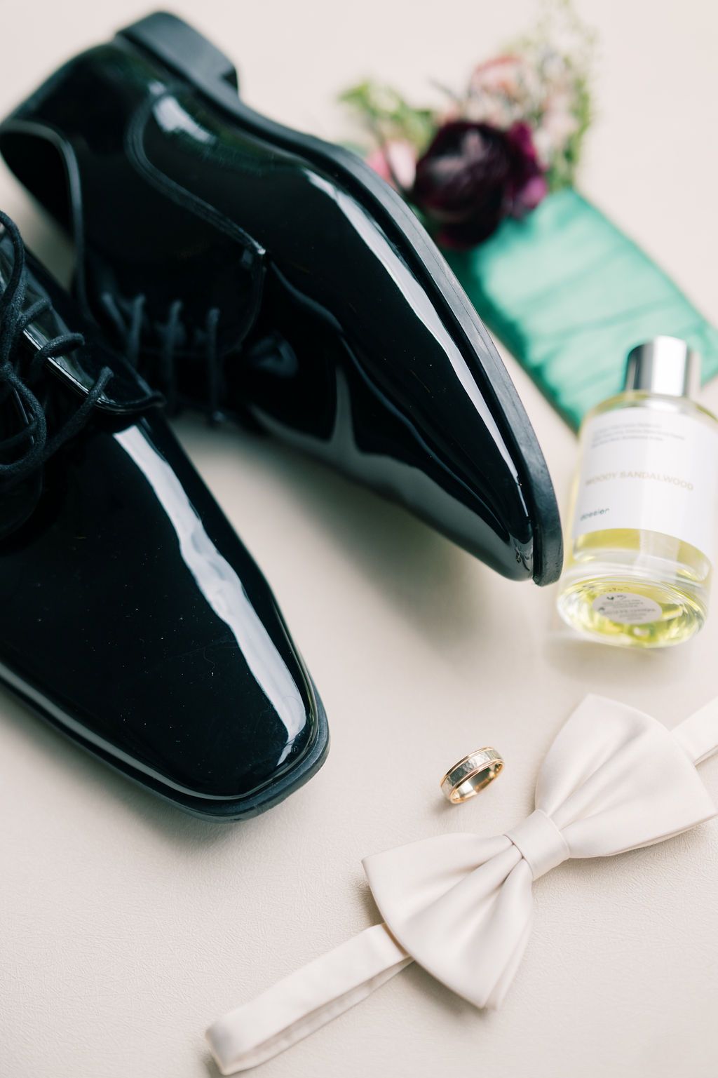 Black patent leather dress shoes with white bow tie, gold wedding rings, cologne bottle, and green pocket square for groom.
