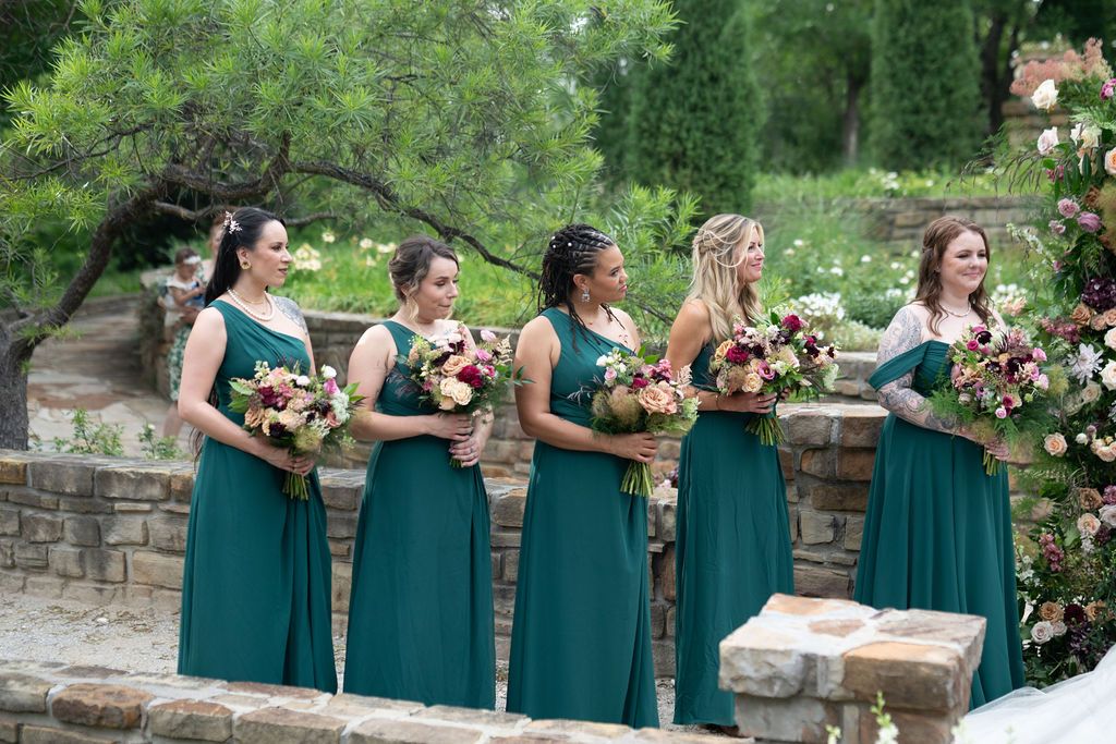 Four bridesmaids in elegant teal green gowns holding floral bouquets stand outdoors at a garden wedding ceremony.