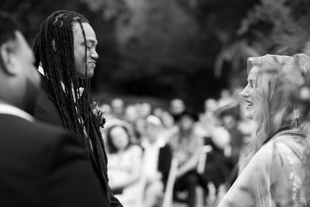 Black and white wedding ceremony photo of a smiling couple exchanging vows outdoors with guests in background.
