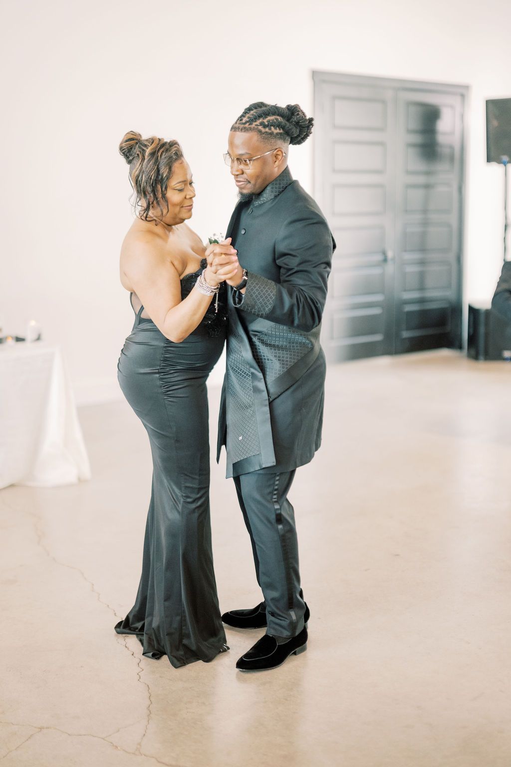 A couple in elegant dark formalwear shares a romantic dance at what appears to be a wedding reception.
