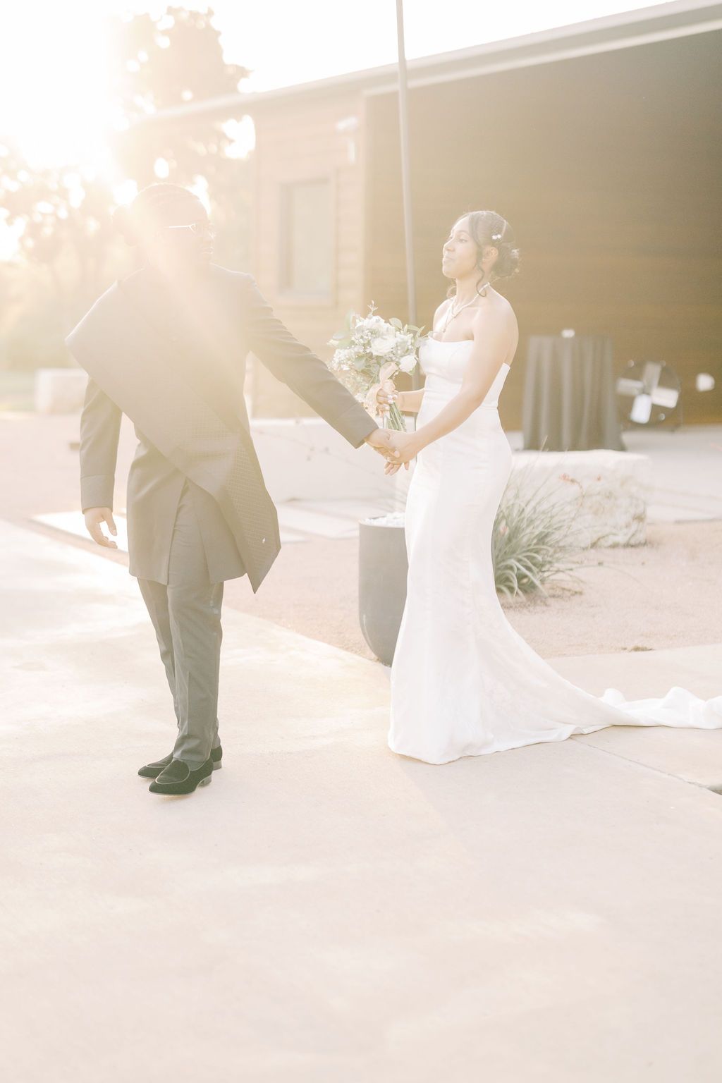 Bride in white gown holds bouquet while groom leads her by hand in golden sunset light outdoors.