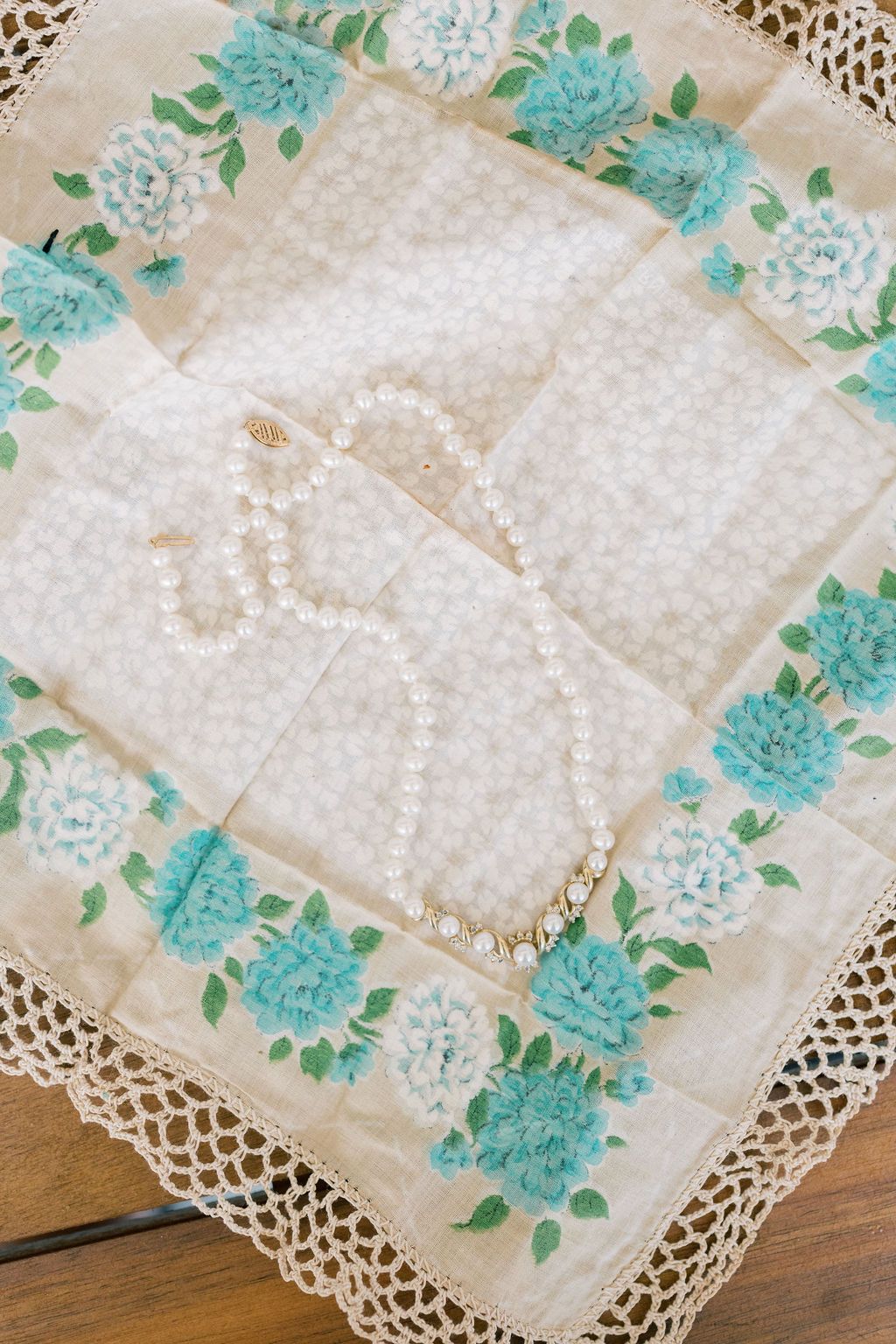 Pearl necklace and bracelet displayed on vintage floral handkerchief with teal flowers and lace trim on wooden surface.