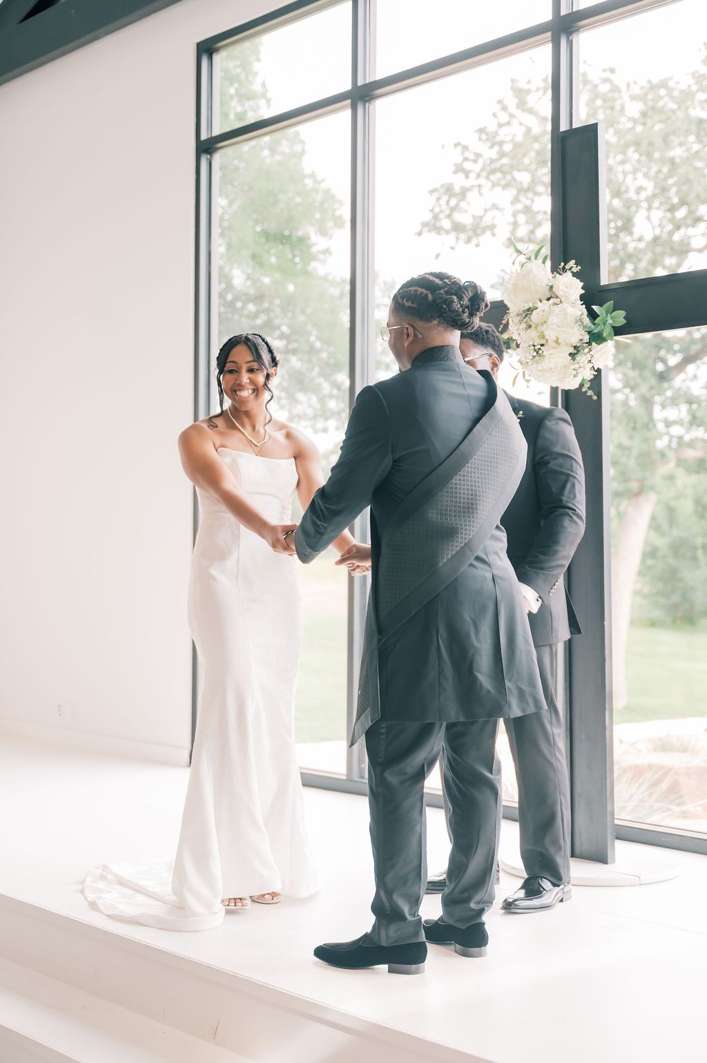 Bride and groom hold hands during wedding ceremony near large windows in a bright modern venue.
