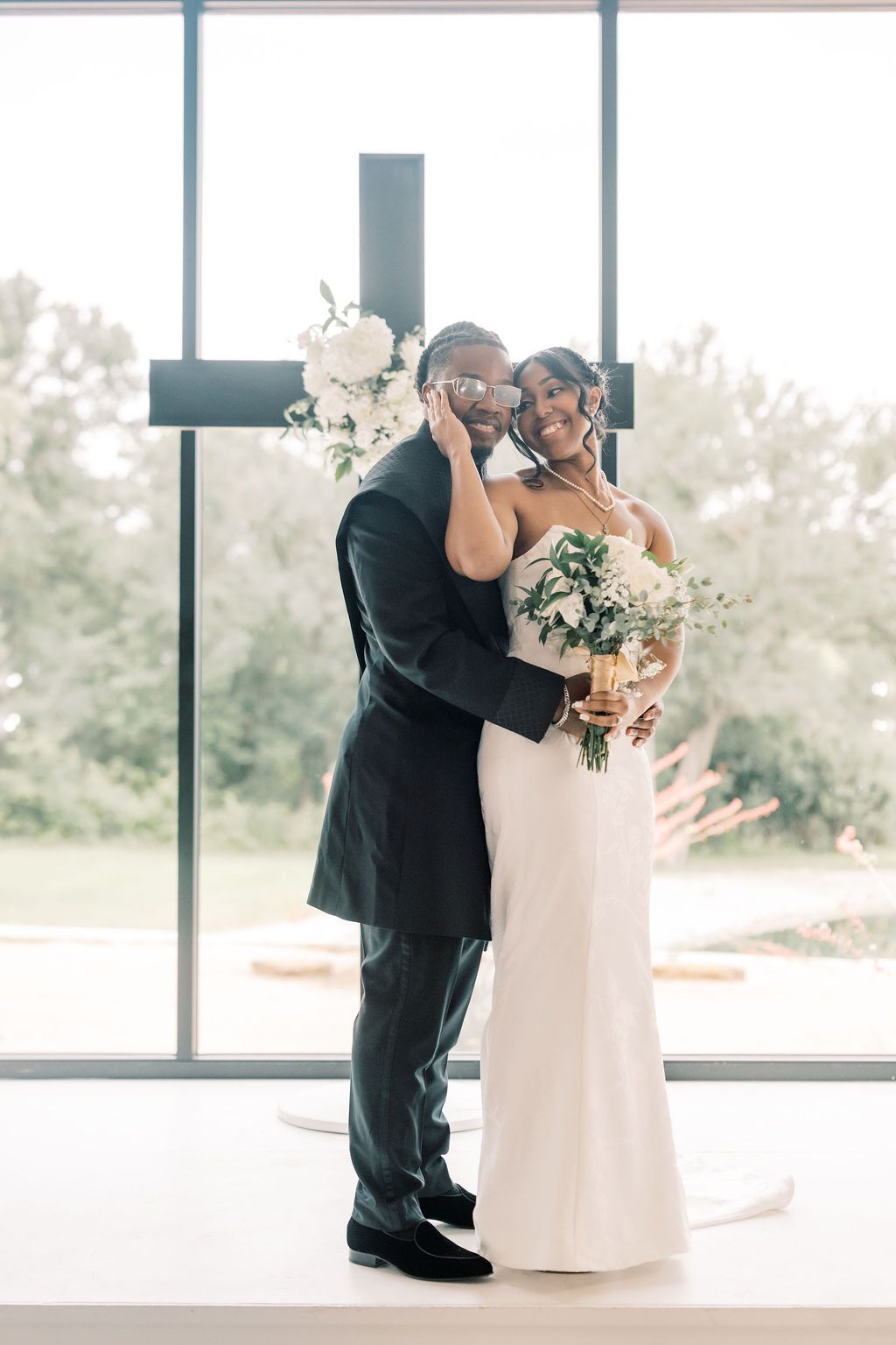 Bride and groom embrace before a cross in a sunlit chapel, bride holding white floral bouquet in elegant gown.