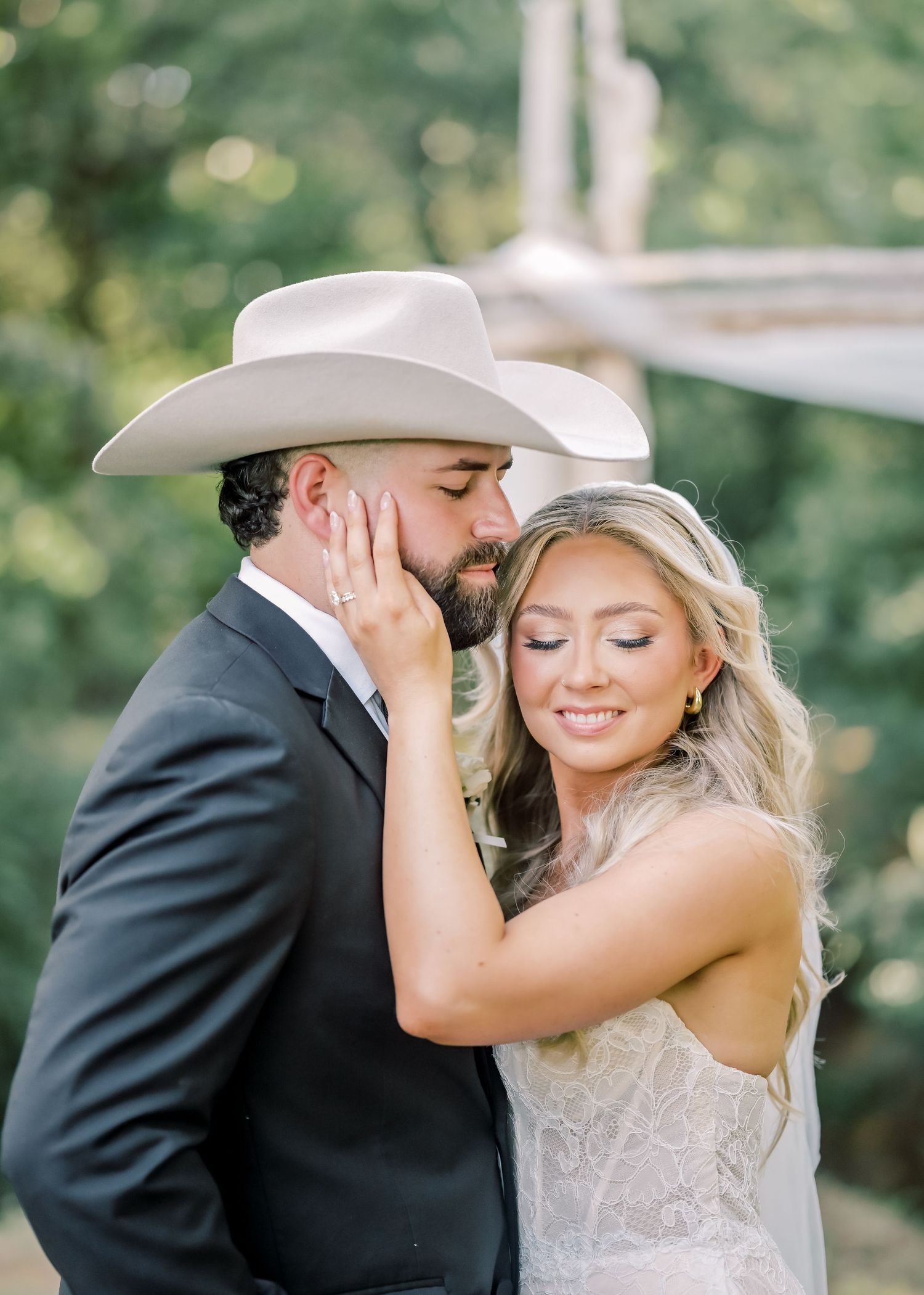 Bearded groom in white cowboy hat tenderly kisses bride's cheek during romantic outdoor wedding photo session.