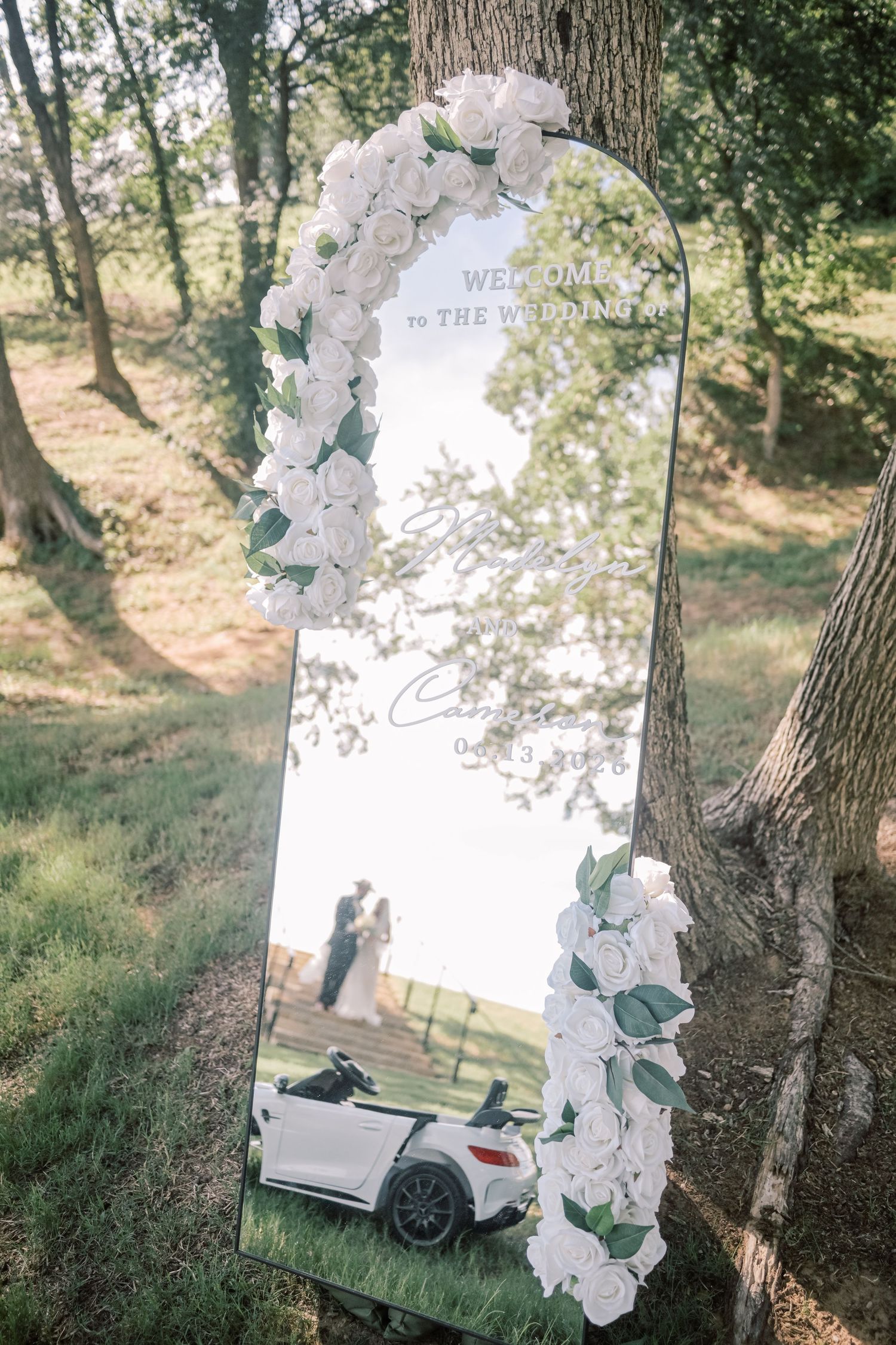 Wedding welcome mirror decorated with white roses reflects couple kissing, leaning against a tree.