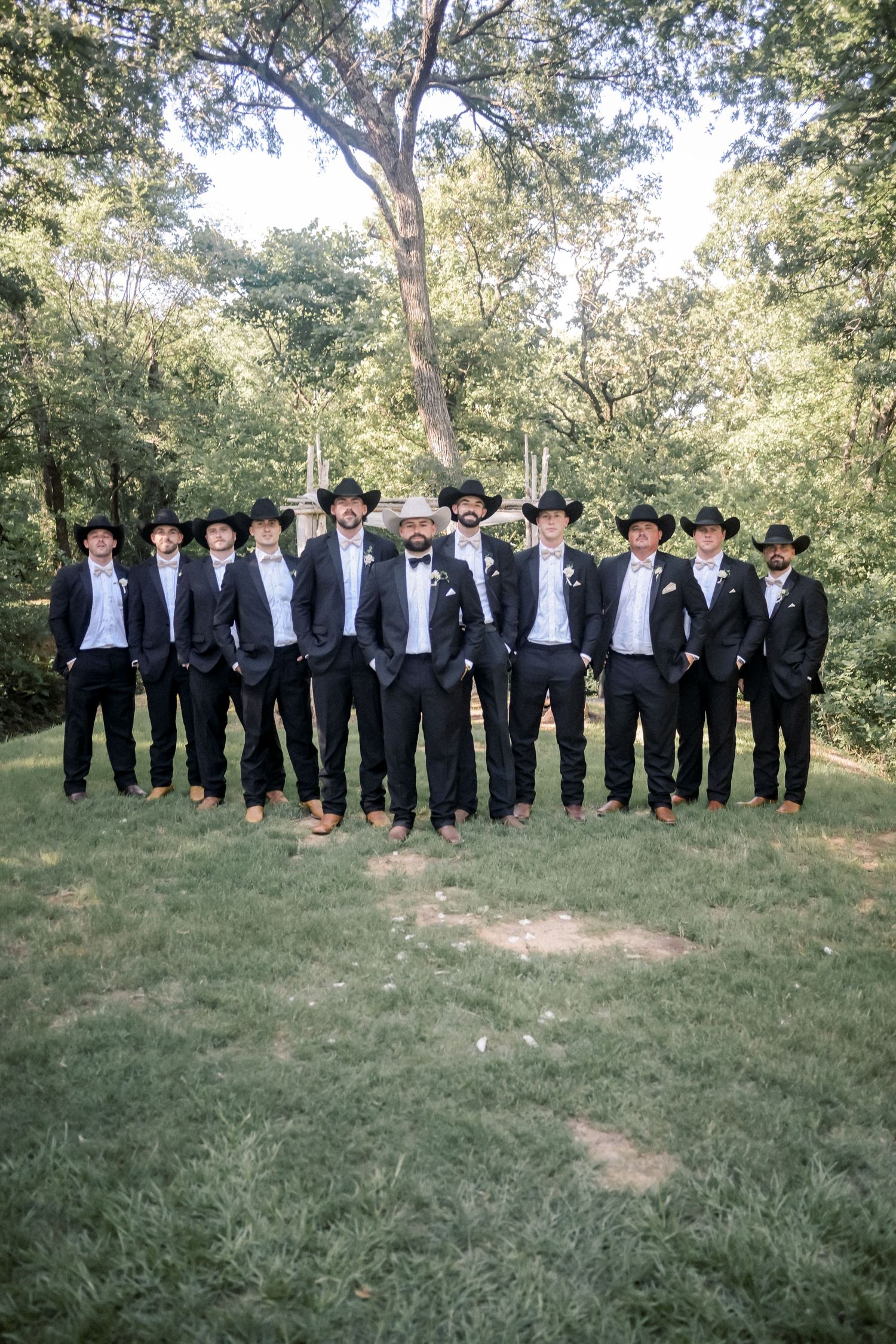 Groomsmen in black suits and cowboy hats pose together on green lawn at outdoor wedding.