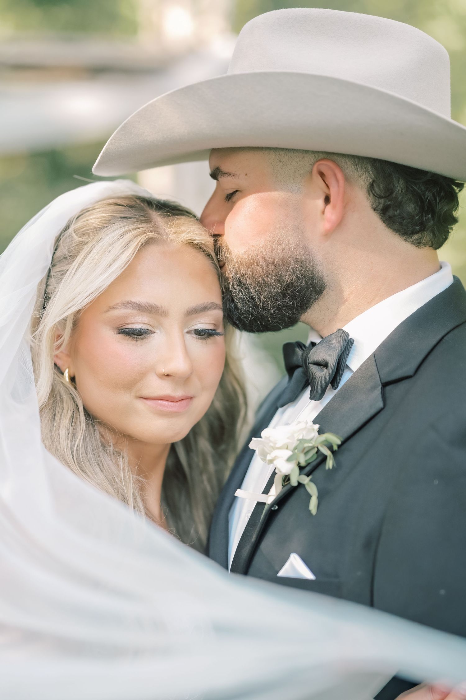 Bearded groom in cowboy hat tenderly kisses blonde bride on forehead during romantic wedding portrait.