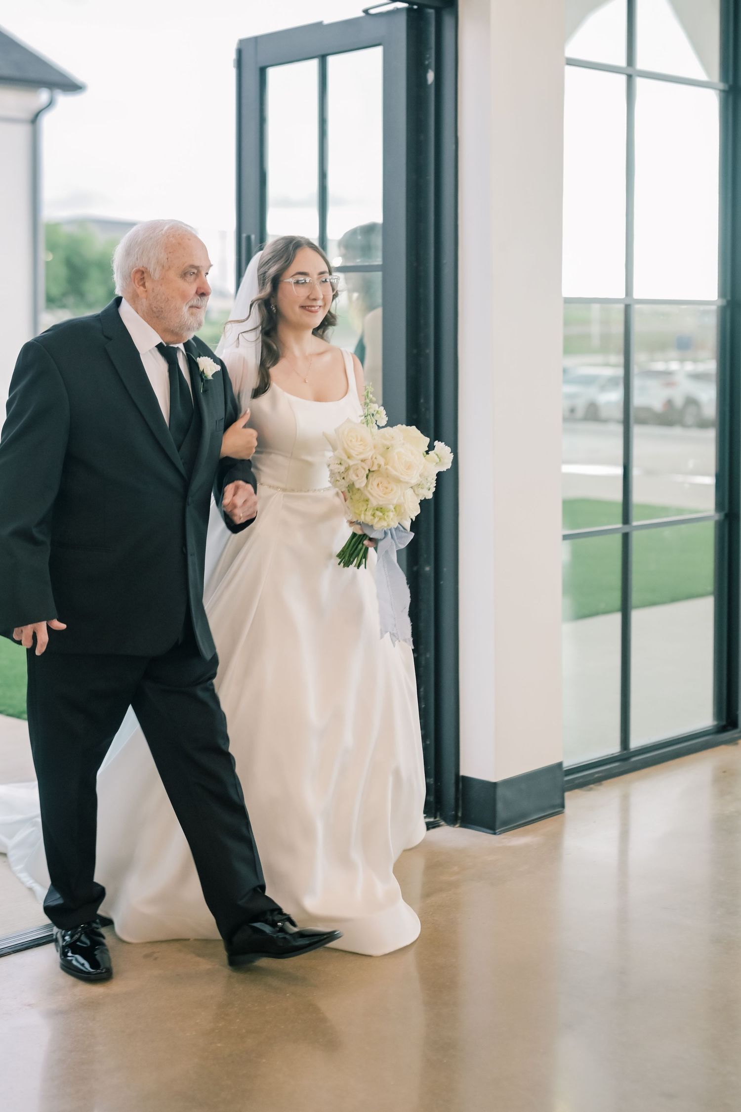 Proud father in black suit escorting smiling bride in white gown through glass doors of wedding venue.