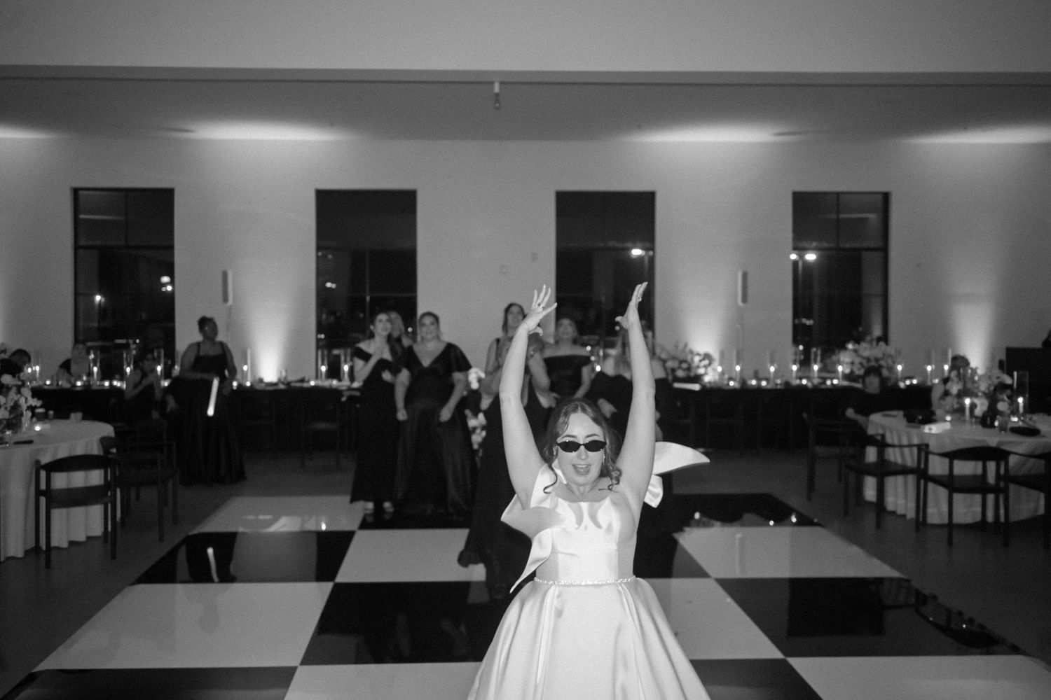 Bride in white gown with sunglasses dancing on checkered floor at wedding reception in black and white.