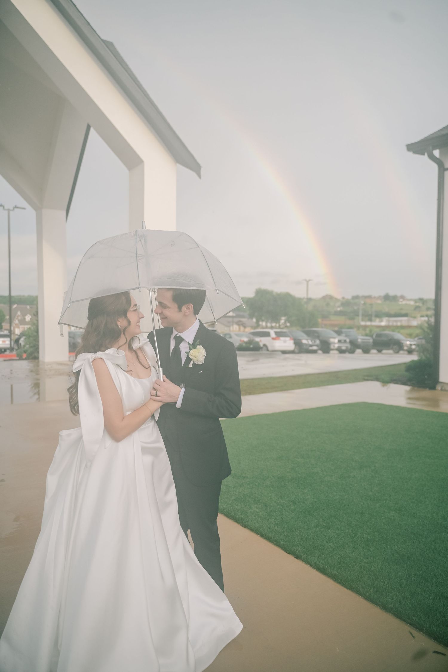 Romantic newlyweds sharing kiss under clear umbrella with stunning double rainbow over wedding venue.