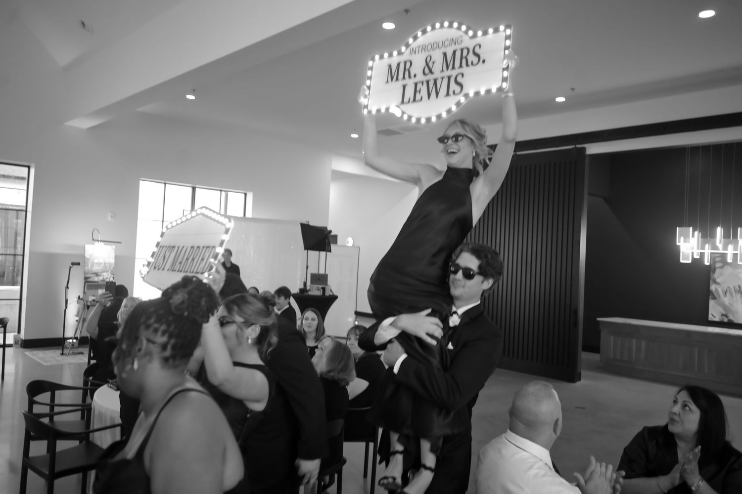 Black and white photo of bride lifted by groom under a Mr. & Mrs. Lewis marquee sign at reception.