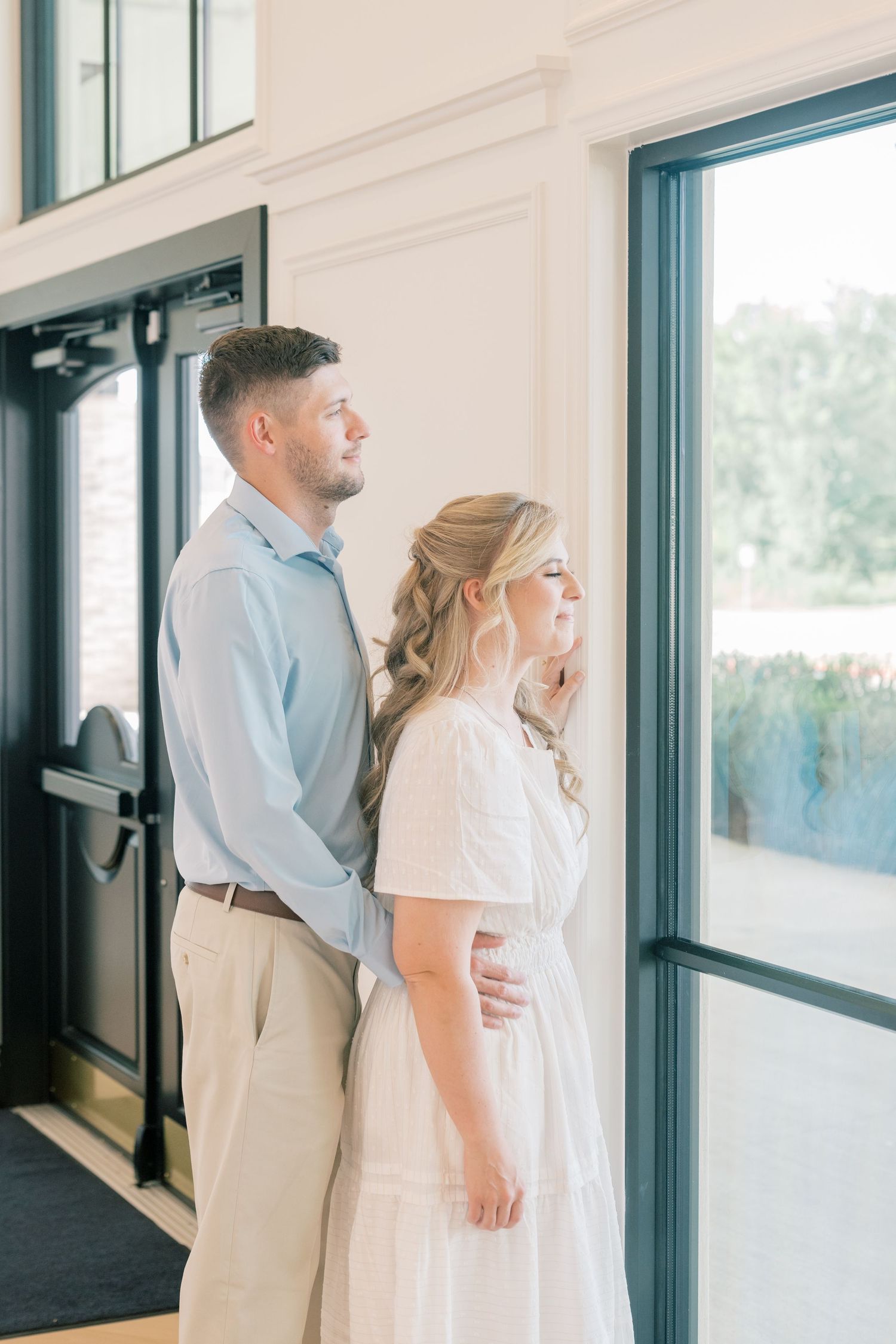 Couple in light blue shirt and white dress standing together near black-framed doors in a bright elegant venue.