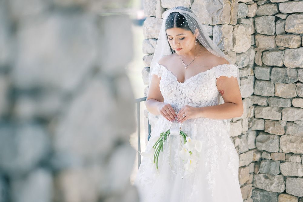An elegant bride in a lace wedding dress poses against a rustic stone wall backdrop.