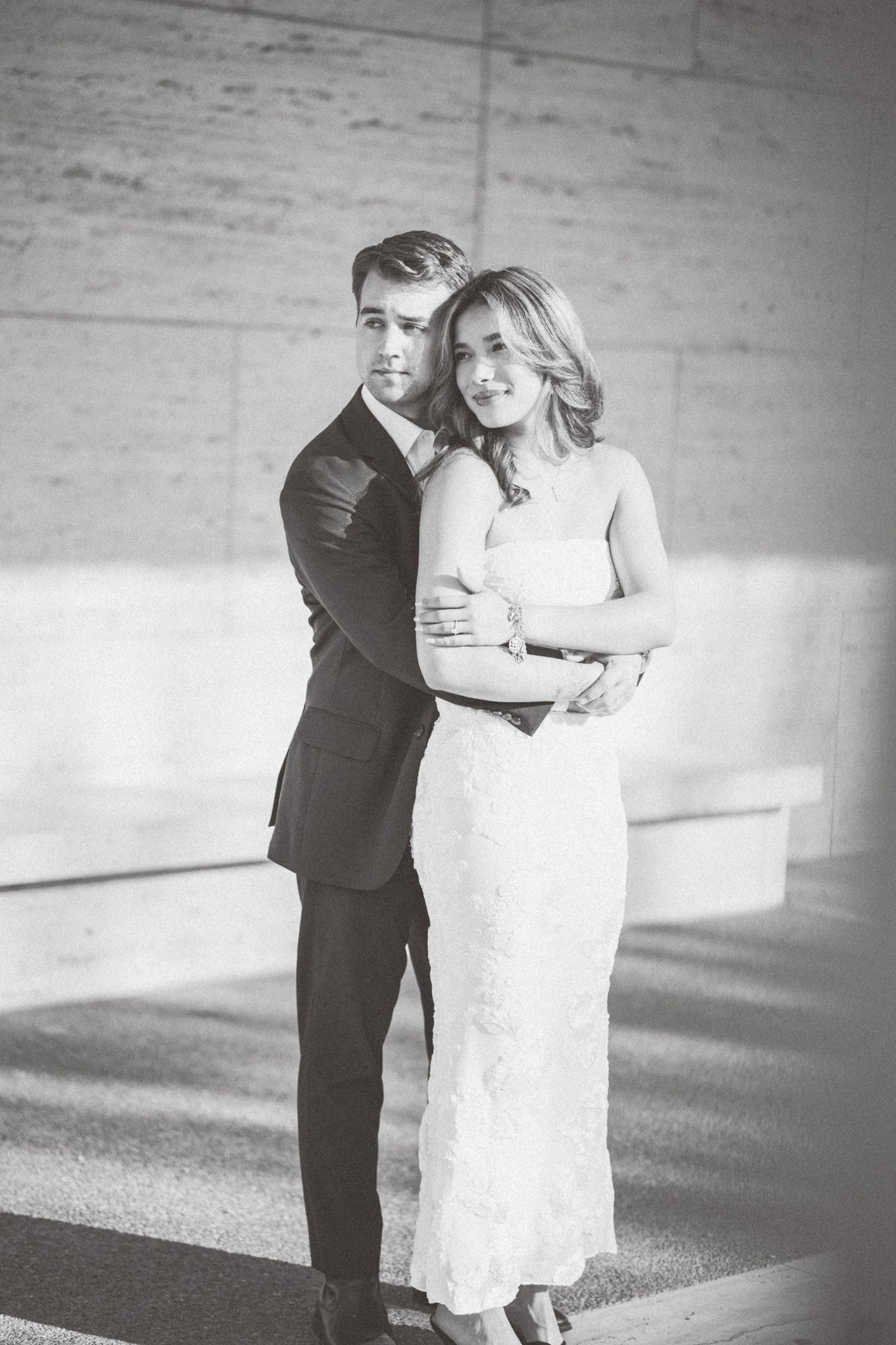A couple in formal attire embraces against a concrete wall in a black and white wedding photo series.