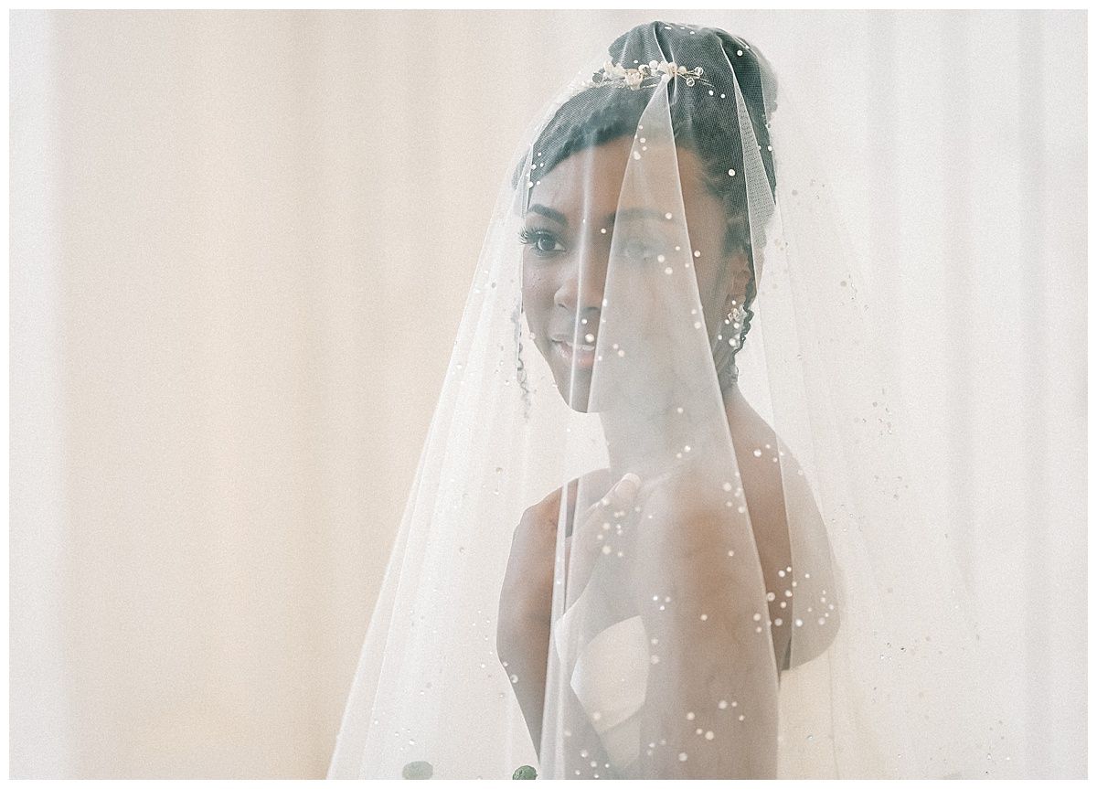 Ethereal bridal portrait through delicate pearl-adorned veil against soft white background.