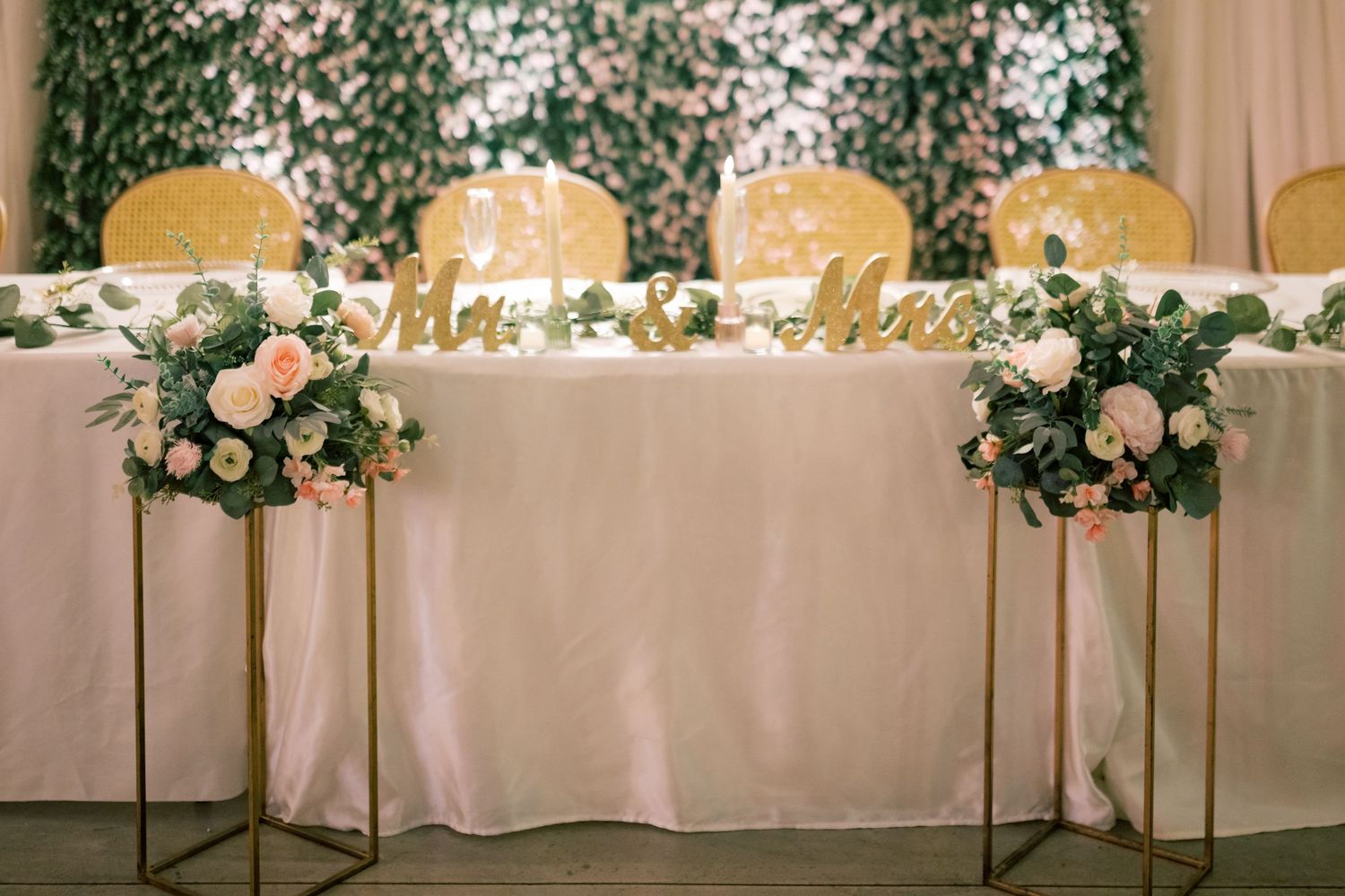 Elegant wedding sweetheart table with gold Mr & Mrs sign, pink floral arrangements, and lush greenery backdrop.