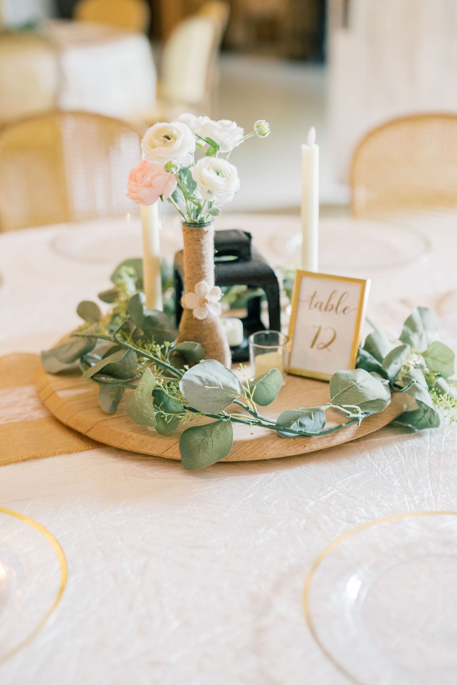 Elegant wedding table centerpiece with white ranunculus, candles, eucalyptus greenery, and gold table number on wooden tray.