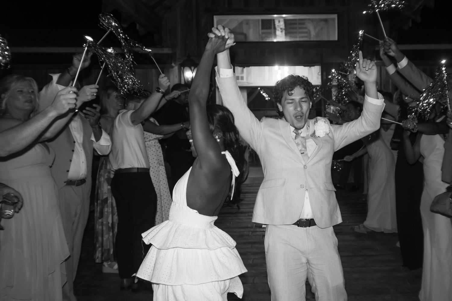 Black and white photo of a joyful couple celebrating at their wedding reception with guests holding sparklers.
