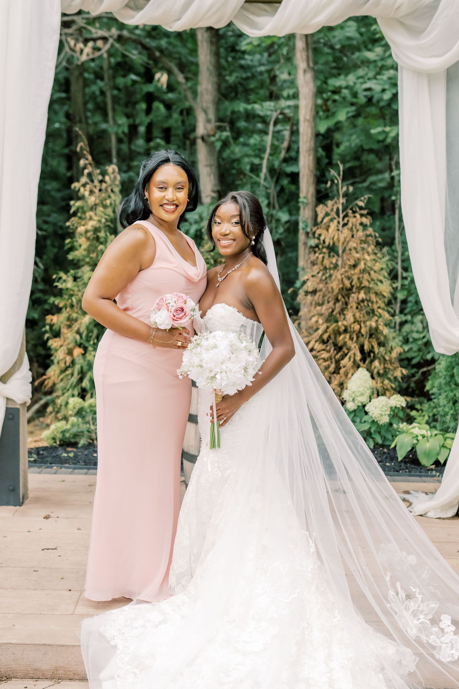 Bride in white lace gown with veil poses with bridesmaid in blush pink dress holding bouquets at outdoor wedding.