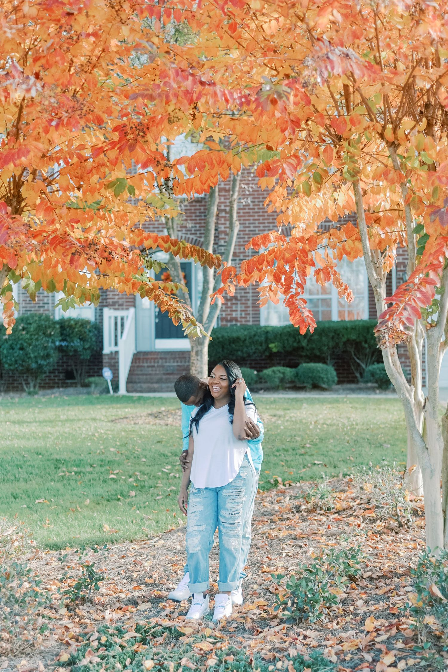Couple embracing under vibrant orange autumn tree leaves in a suburban yard during fall season.