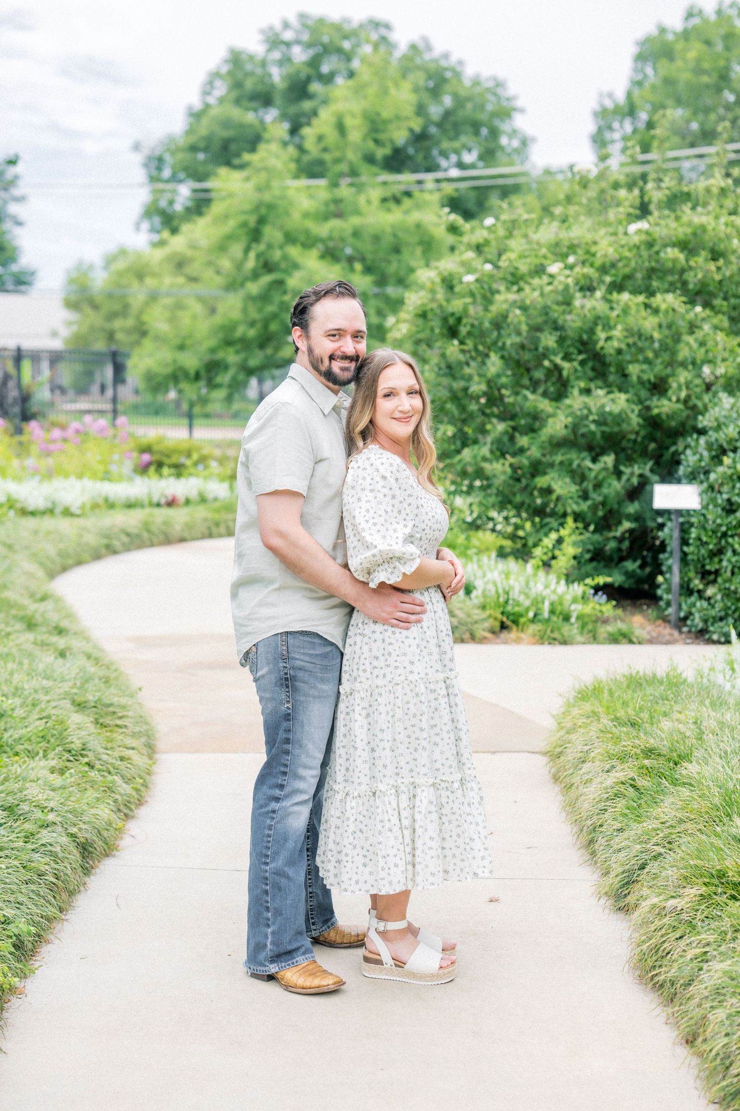 Couple embracing on a garden path, woman in floral dress and man in gray shirt and jeans, surrounded by lush greenery.
