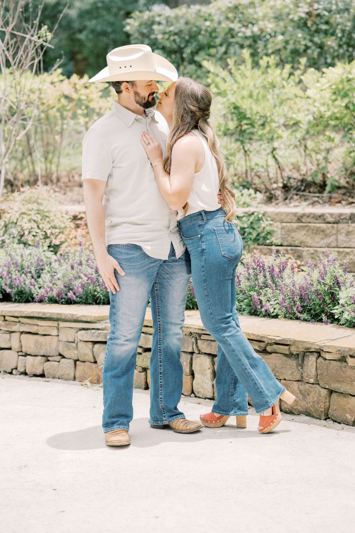 A couple shares a kiss outdoors, man wearing a cowboy hat and jeans, woman in white top and flared jeans.