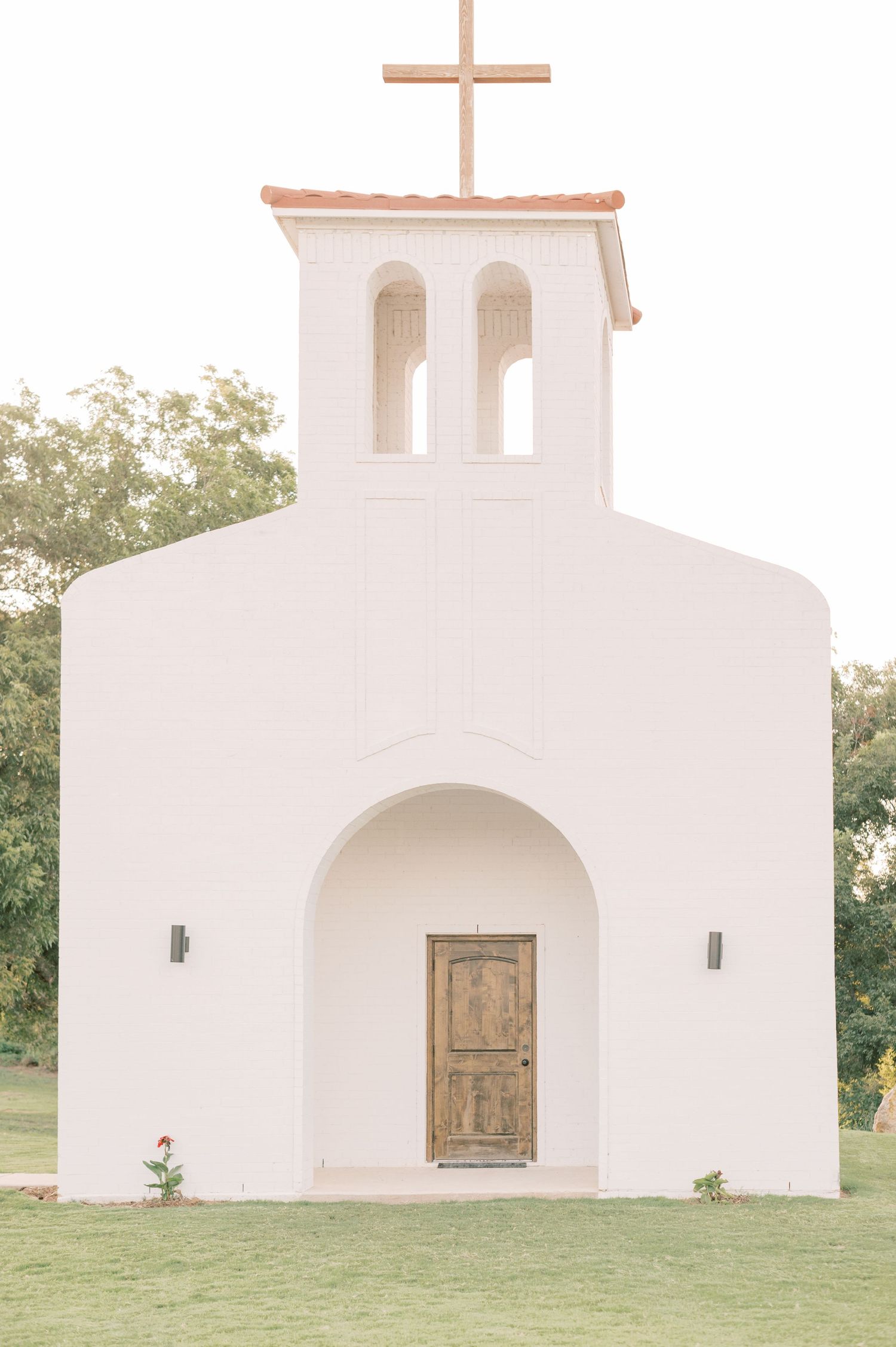 White stucco church with wooden doors and arched windows sits peacefully on a grassy landscape.