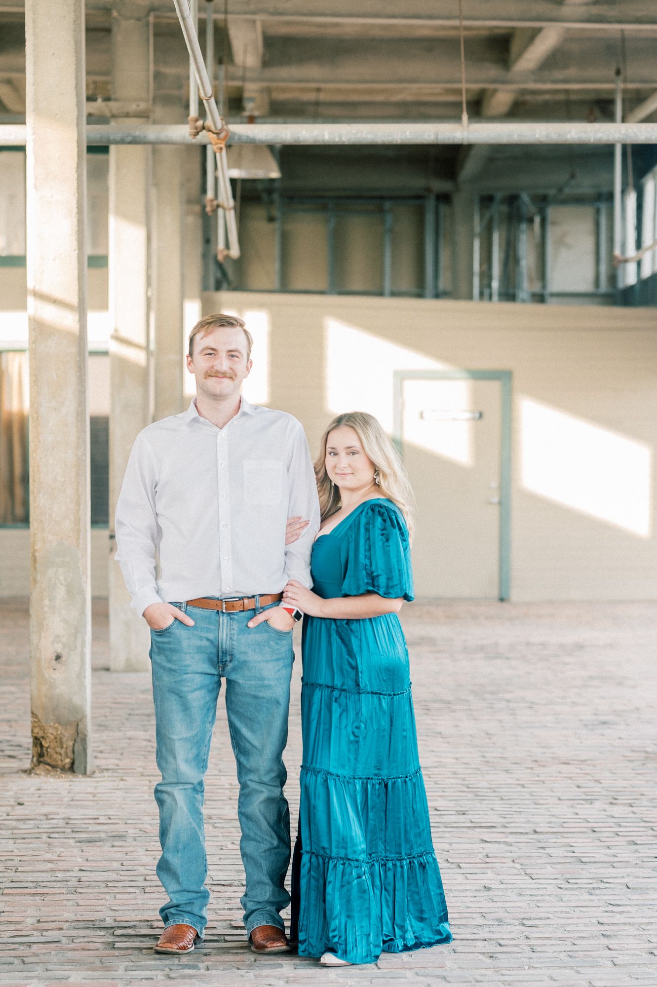A couple poses together in an industrial warehouse setting with sunlight streaming through windows.