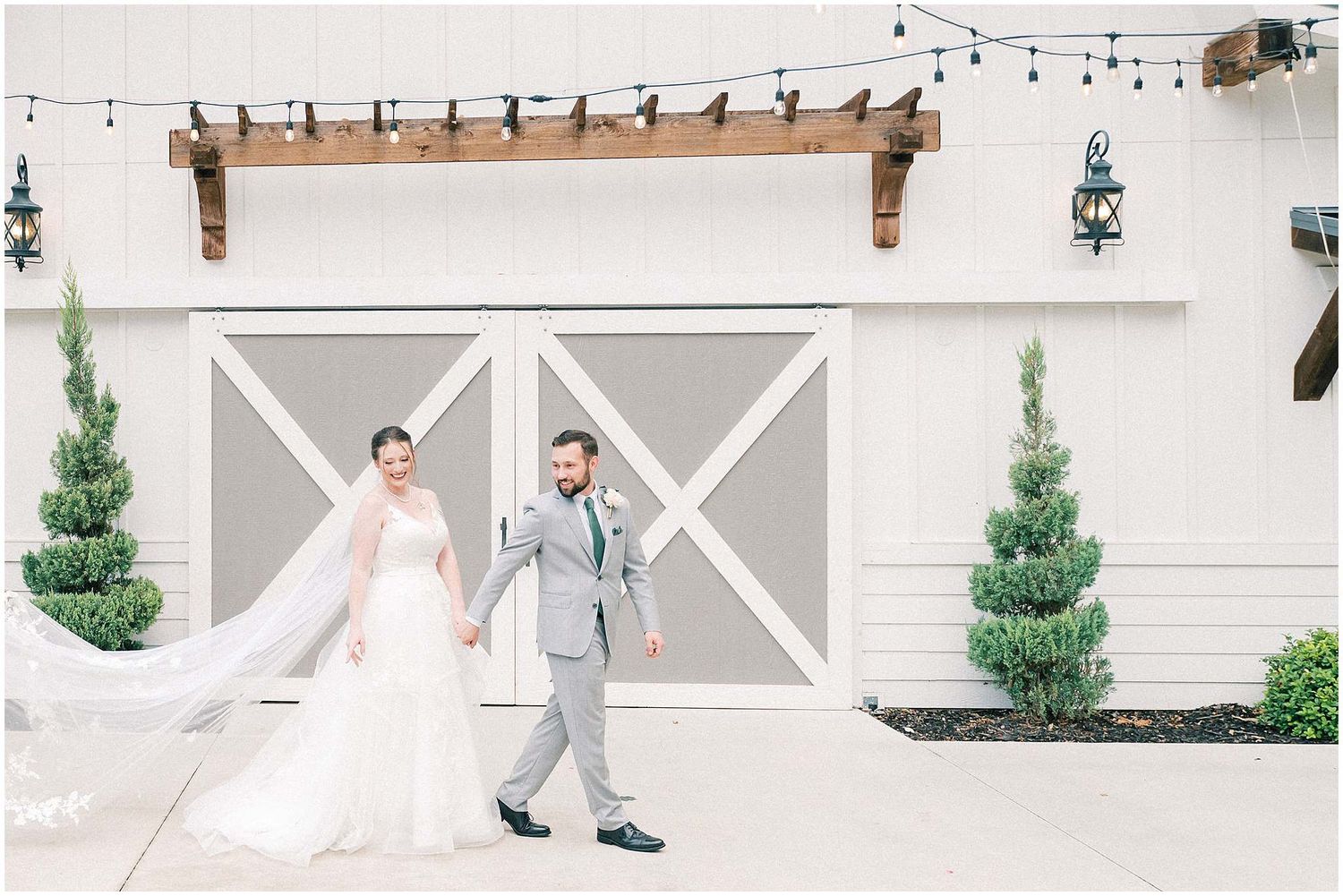 A wedding couple walks along a white barn-style building with decorative evergreen trees and cross-patterned walls.