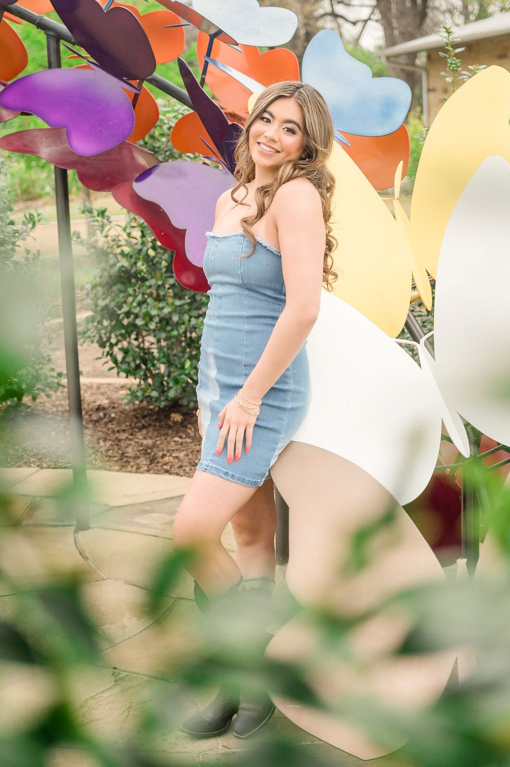 Young woman in denim dress and boots posing among colorful giant flower sculptures in an outdoor garden setting.