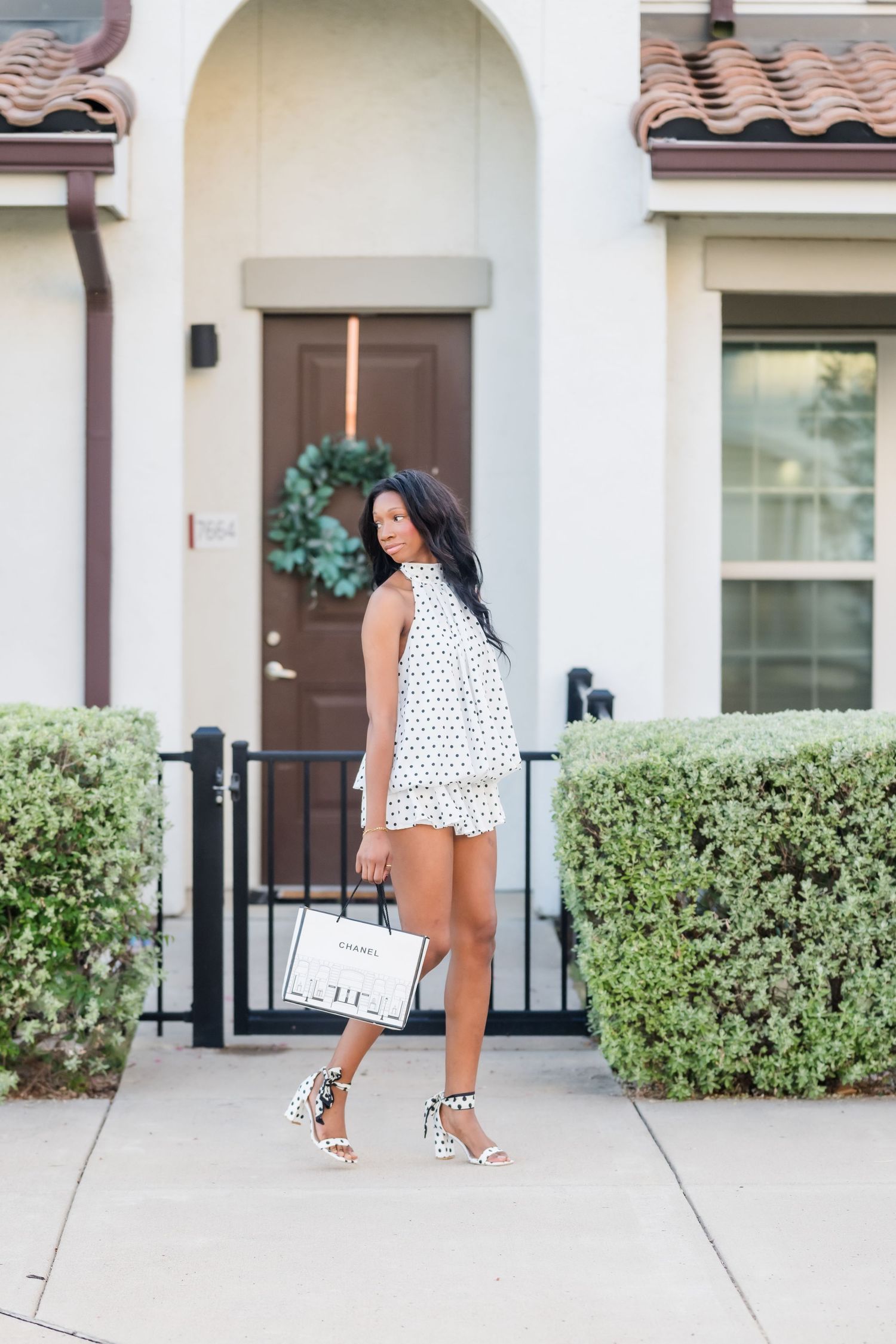 Woman in white polka dot romper and leopard heels poses on sidewalk outside a home with a wreath on the door.