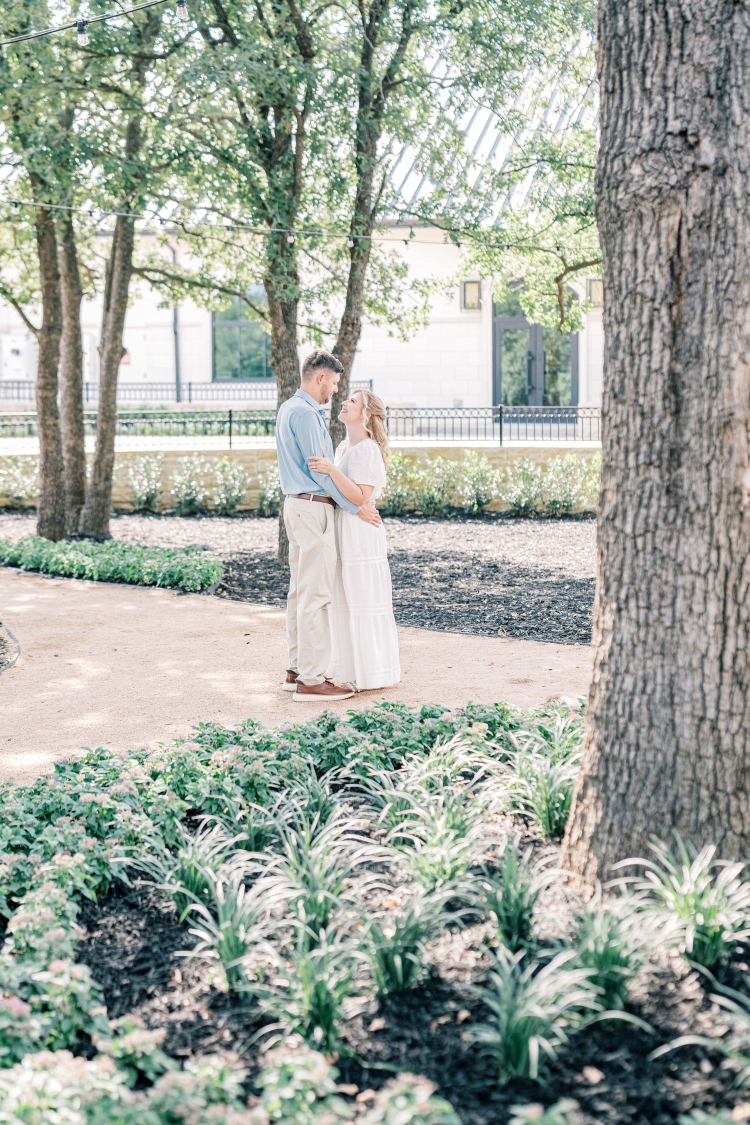 Couple stands together on a garden path surrounded by lush green trees and flowering plants.