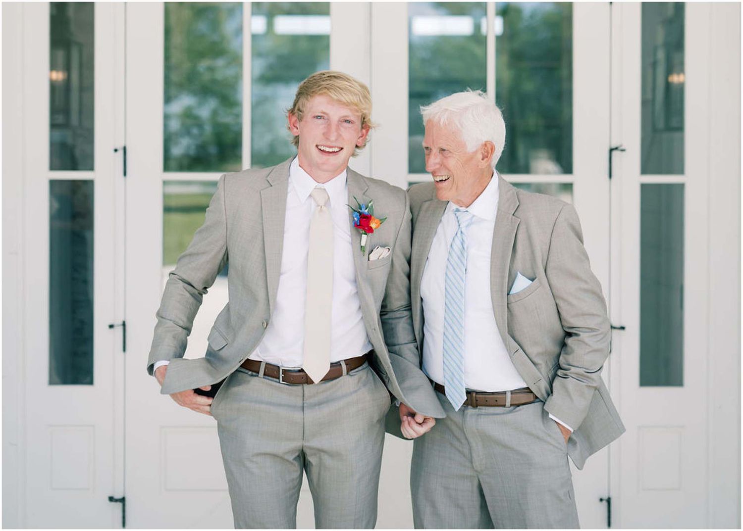 Two men in matching gray suits with boutonnieres smile together at a wedding ceremony.