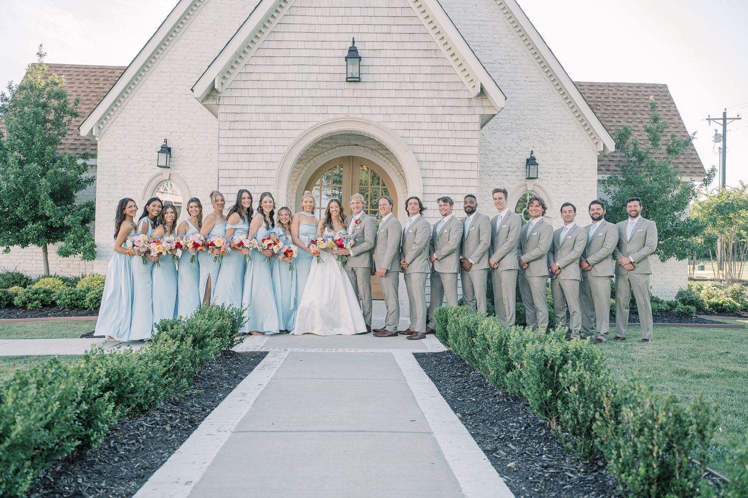 Wedding party posing outside stone chapel, bridesmaids in sage green gowns and groomsmen in tan suits.