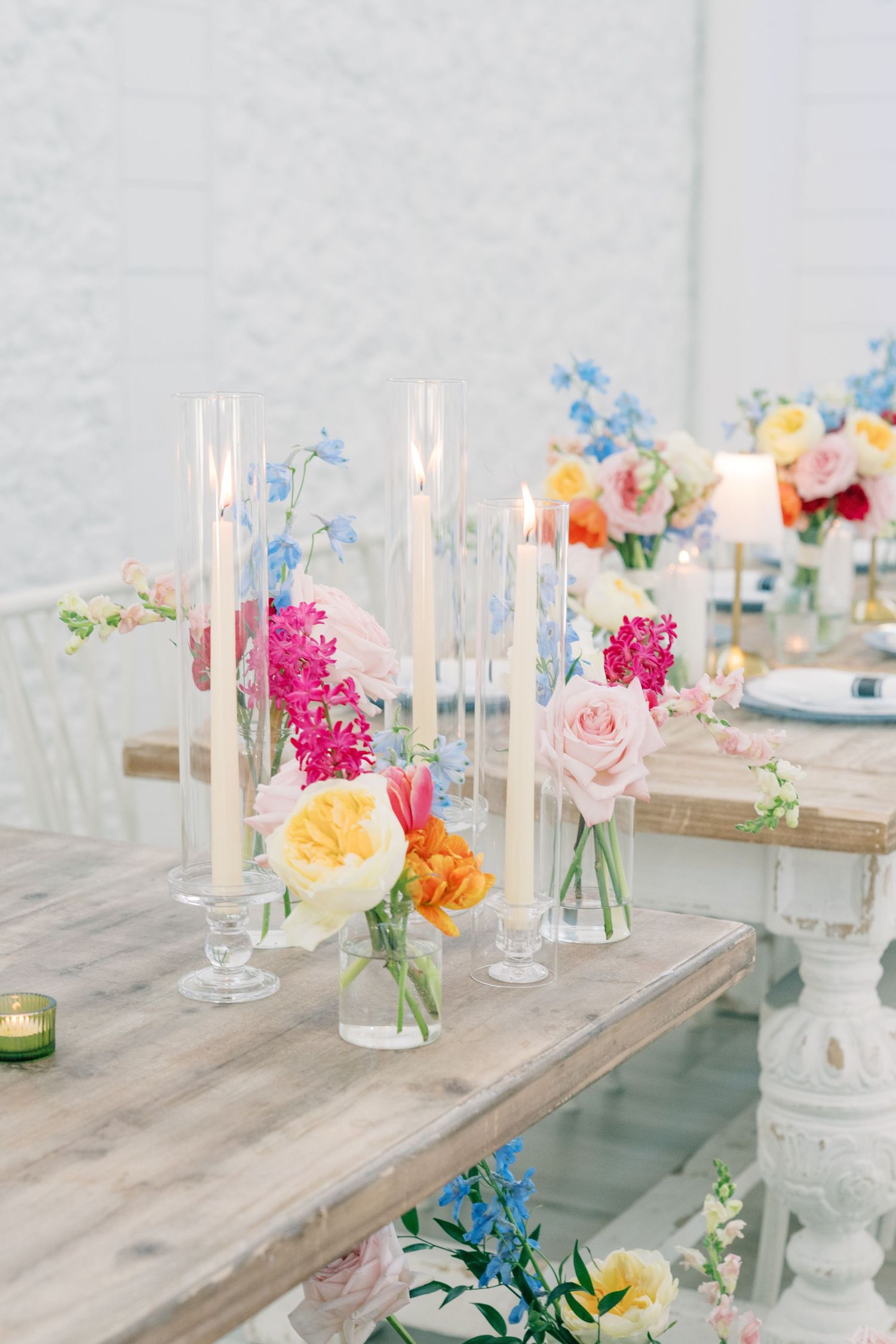 Elegant wedding table with tall glass candle holders and colorful floral arrangements in pink, yellow, and blue hues.