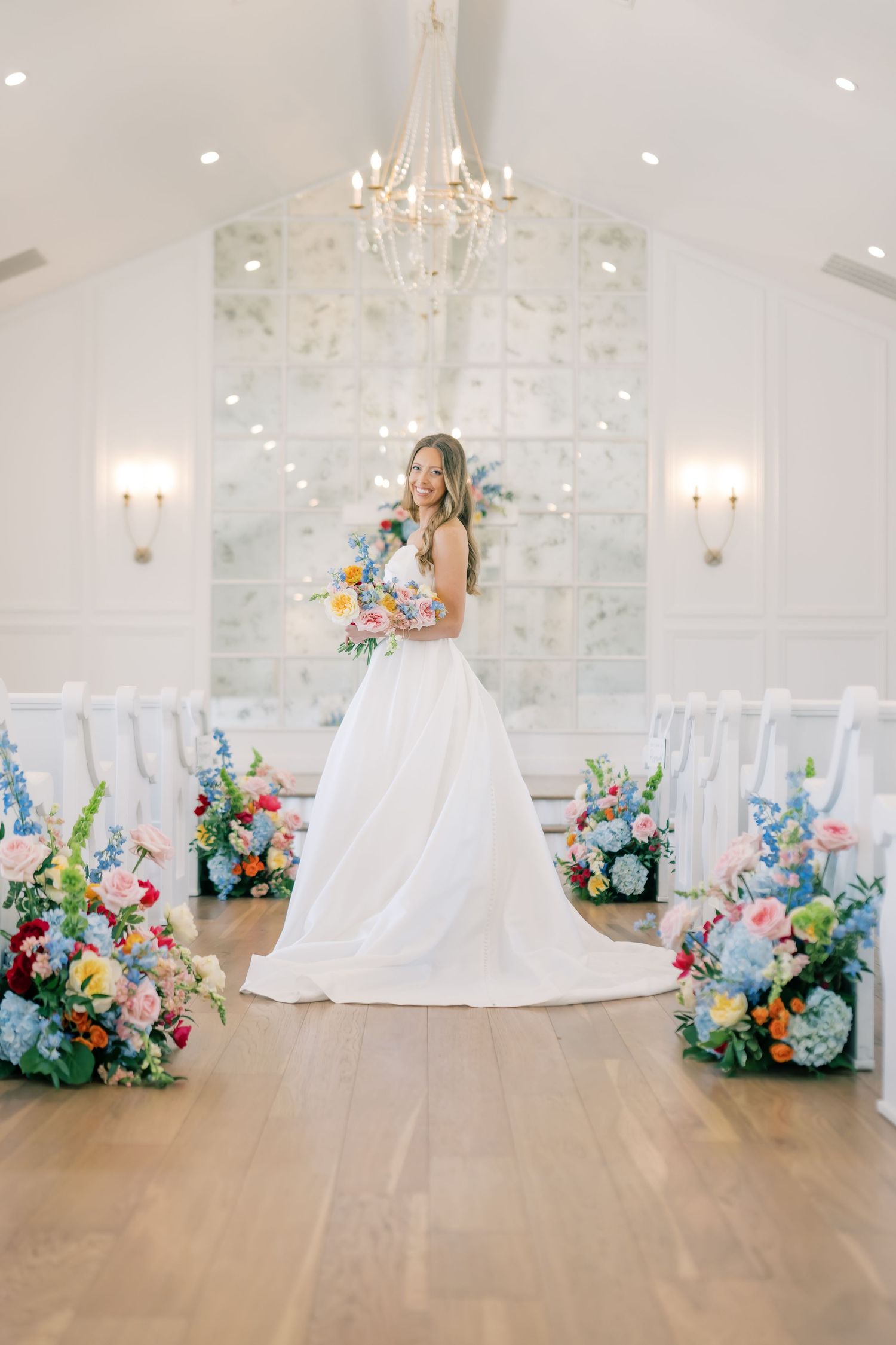 Bride in white ball gown holds colorful bouquet in bright chapel with floral arrangements lining the aisle.